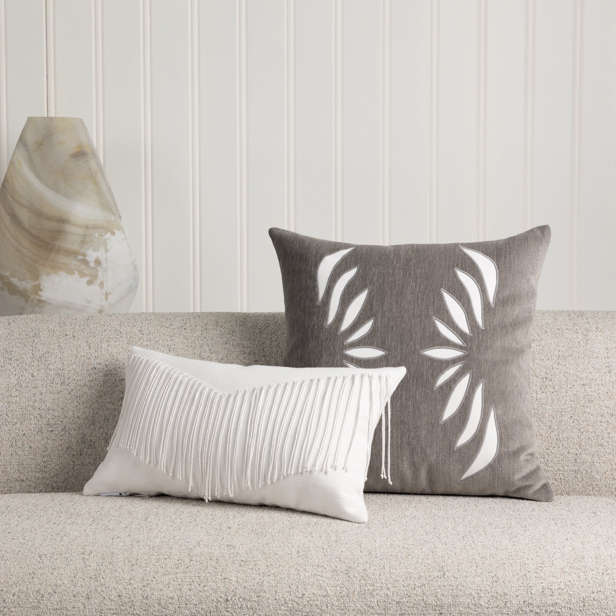 Indoor image of gray velour pillow with white abstract cutouts on ivory sofa with white lumbar with fringe detailing