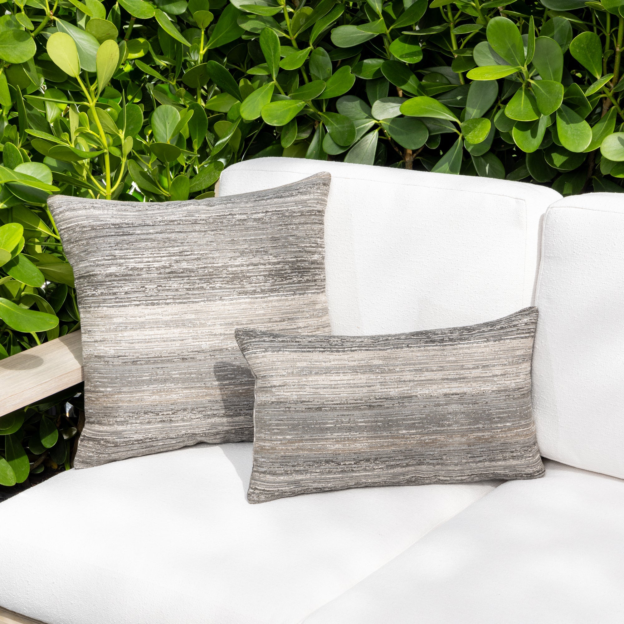 Outdoor image of pillows in Two gray Elaine Smith pillows on a white outdoor sofa with green foliage in the background