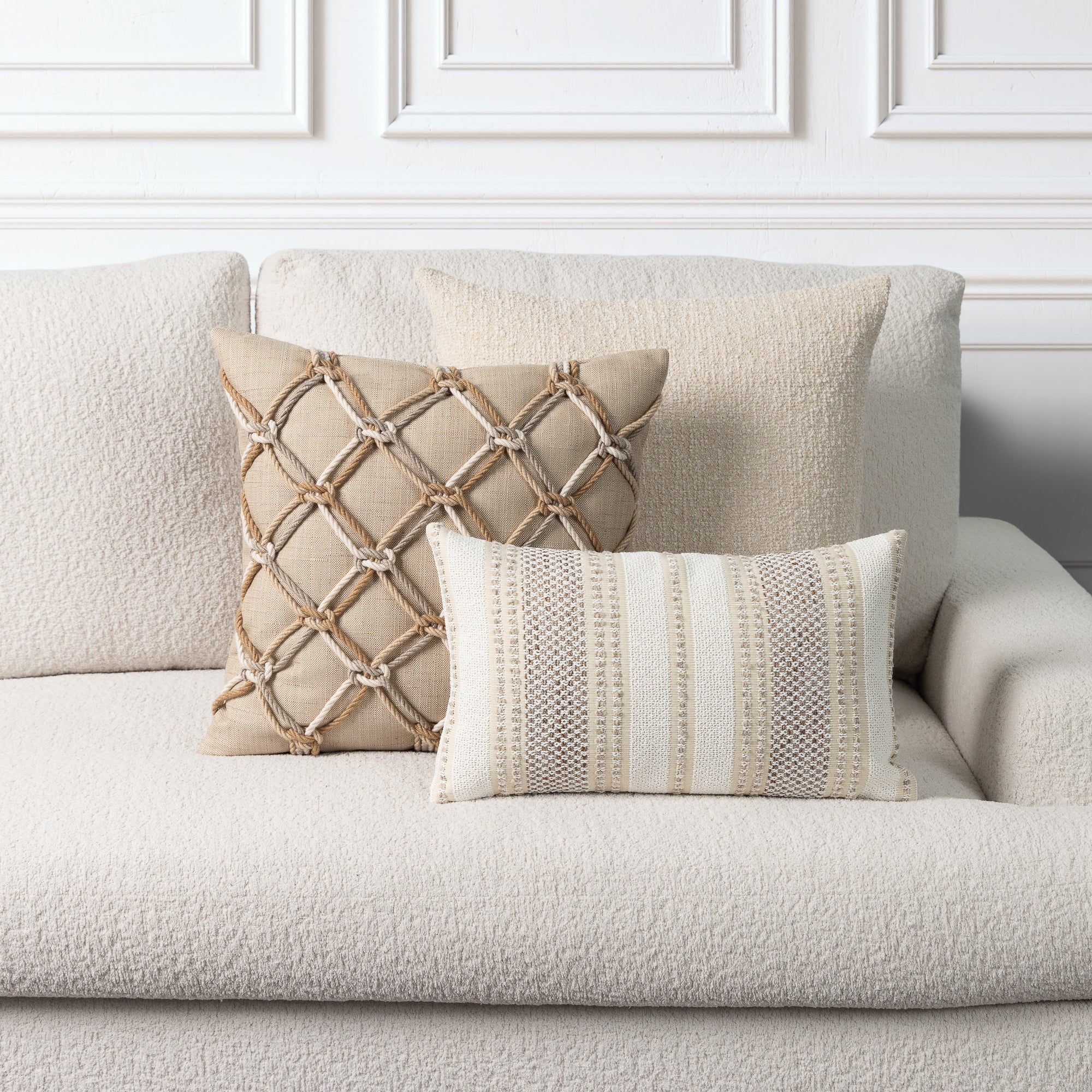 Two decorative throw pillows on a textured cream boucle sofa against a white paneled wall. One pillow is beige with a rope lattice pattern, and the other is cream and beige with vertical woven stripes.
