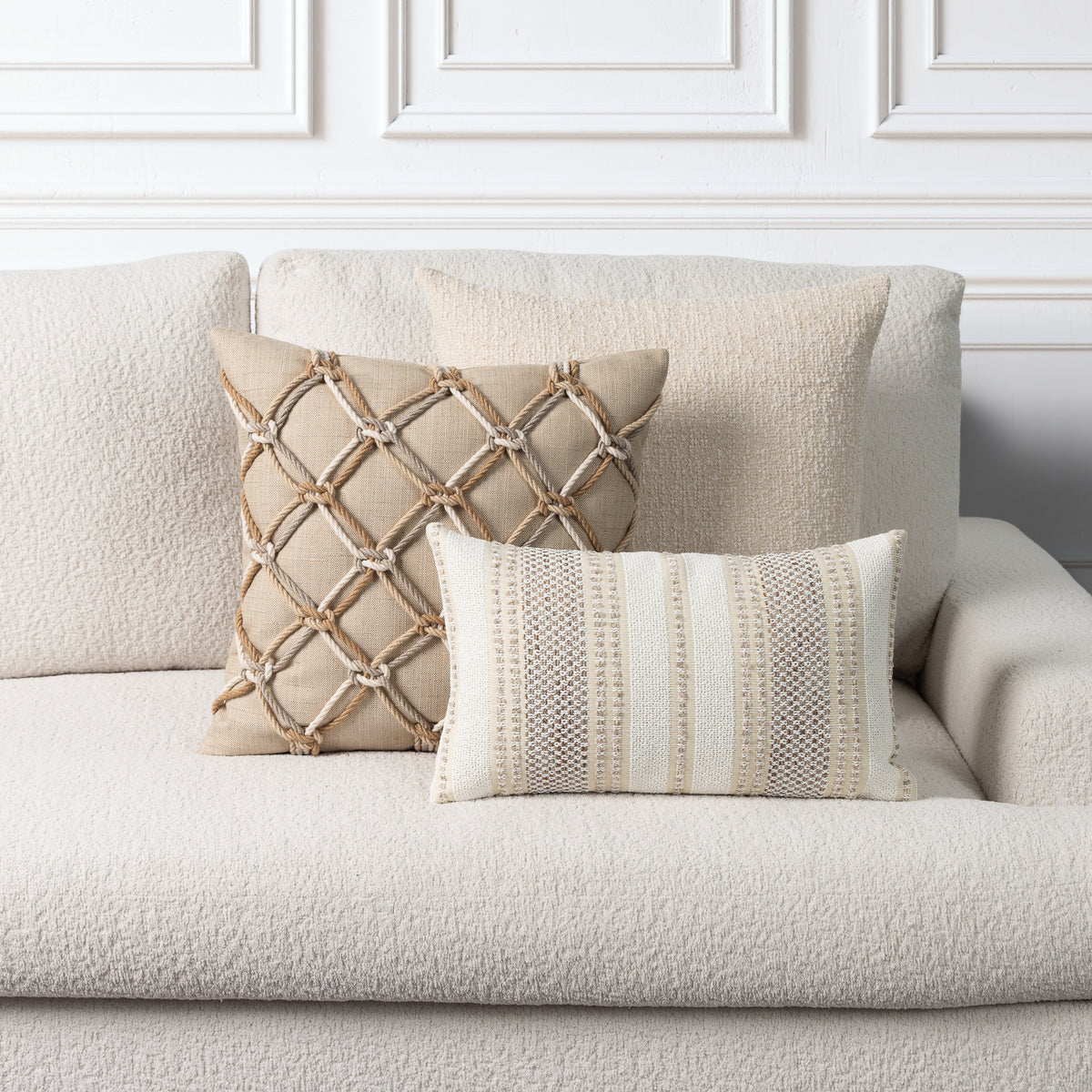 Two decorative throw pillows on a textured cream boucle sofa against a white paneled wall. One pillow is beige with a rope lattice pattern, and the other is cream and beige with vertical woven stripes.