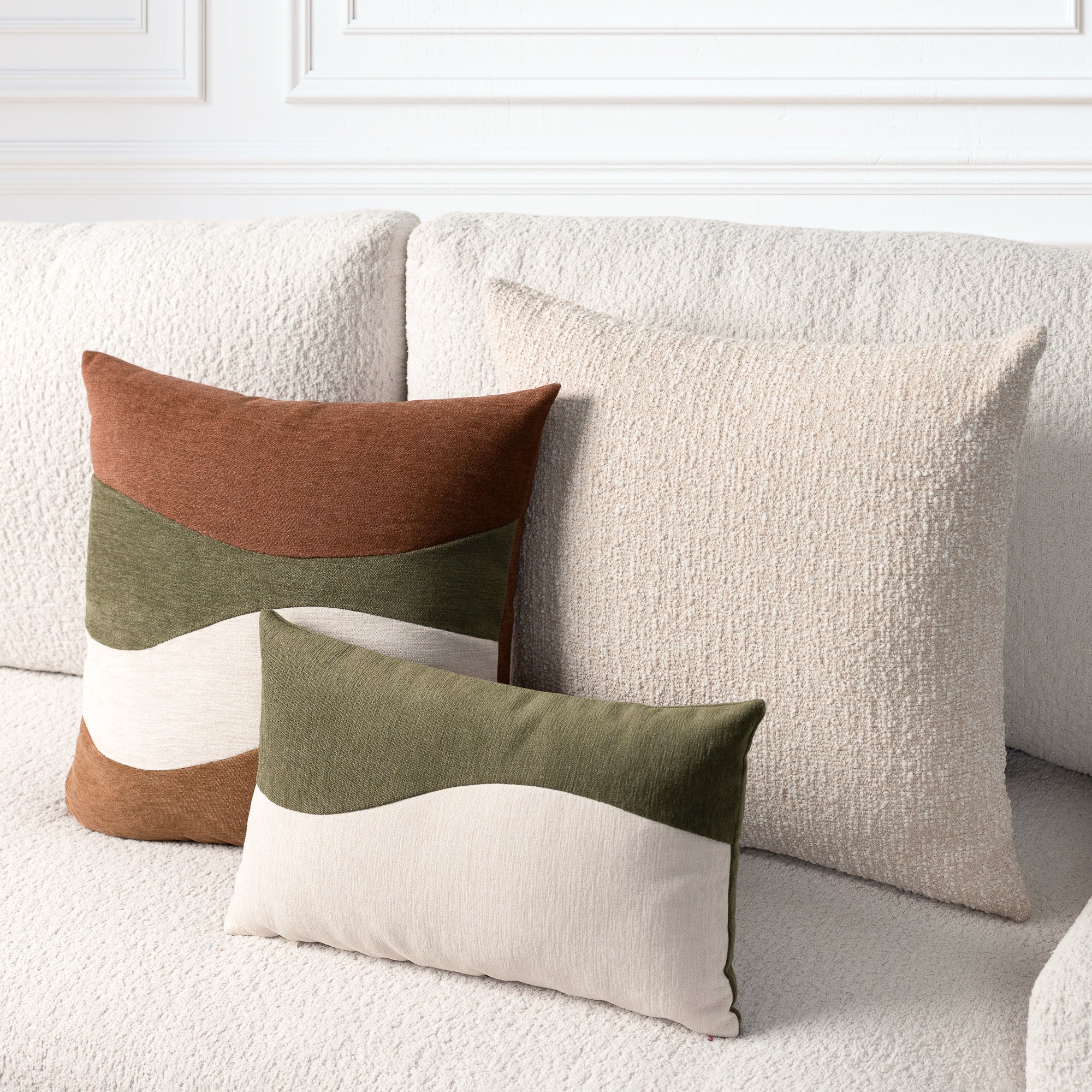 Three decorative throw pillows arranged on a textured cream sofa in a bright living room setting. The set includes a large cream pillow, a smaller cream pillow, and a rust-brown and olive green color-blocked pillow.