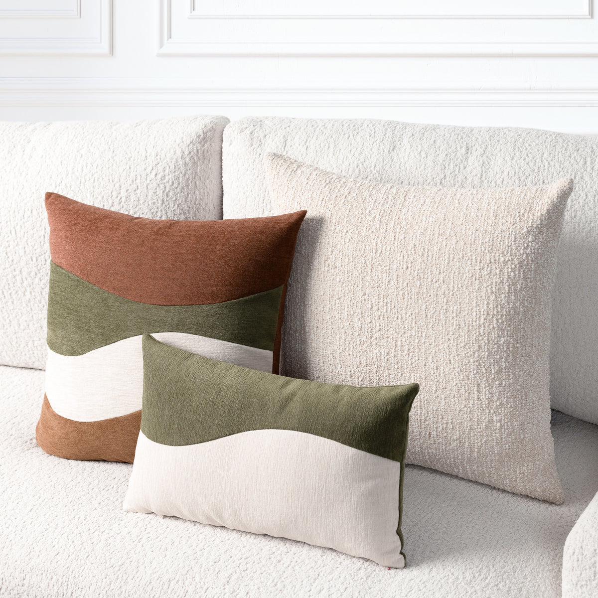 Three decorative throw pillows arranged on a textured cream sofa in a bright living room setting. The set includes a large cream pillow, a smaller cream pillow, and a rust-brown and olive green color-blocked pillow.