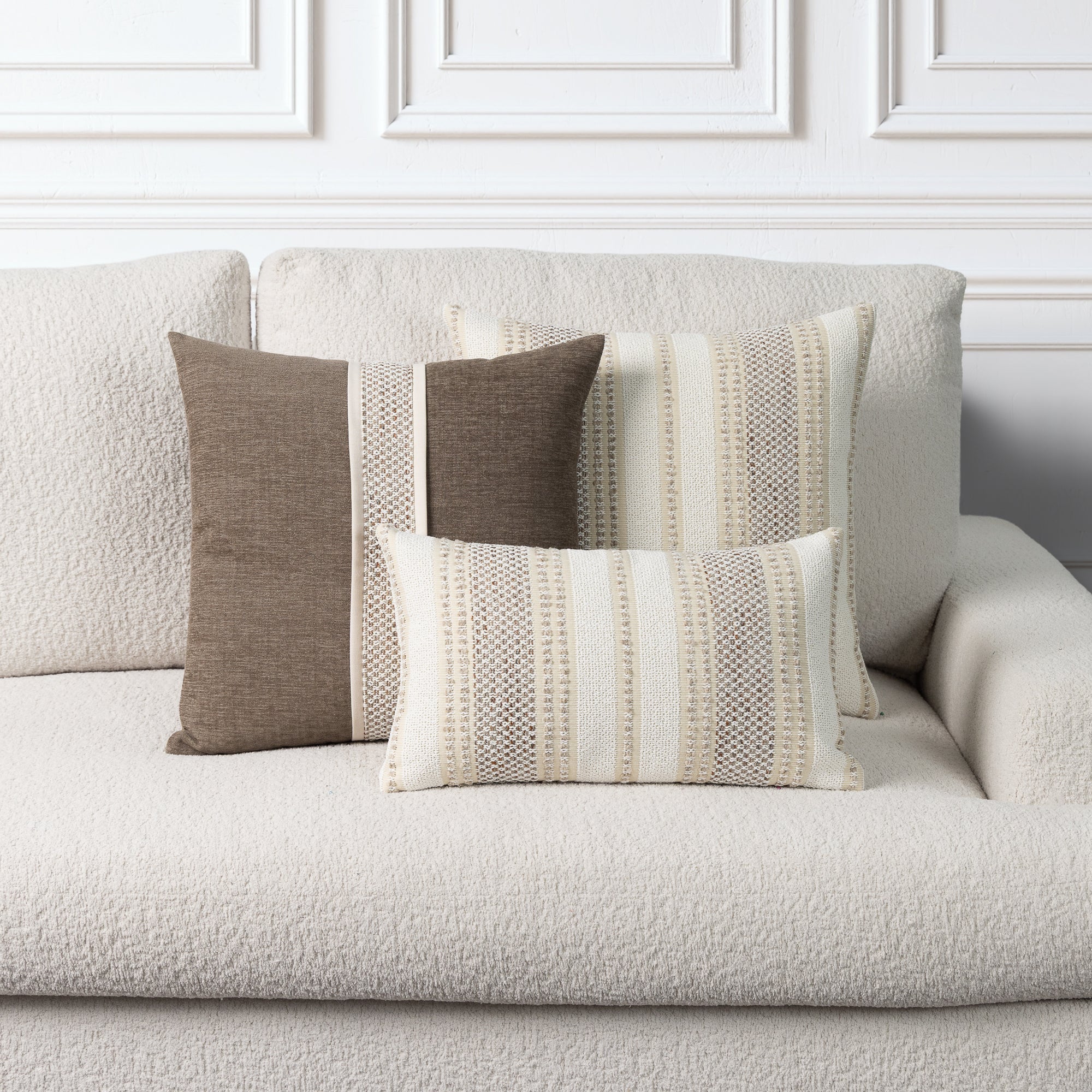 Three decorative throw pillows in neutral tones of brown, beige, and cream, styled on a textured off-white boucle sofa against a white paneled wall.