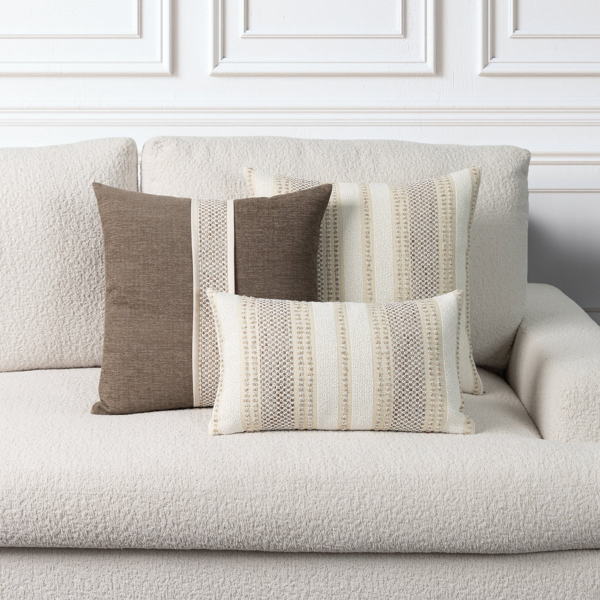 Three decorative throw pillows in neutral tones of brown, beige, and cream, styled on a textured off-white boucle sofa against a white paneled wall.