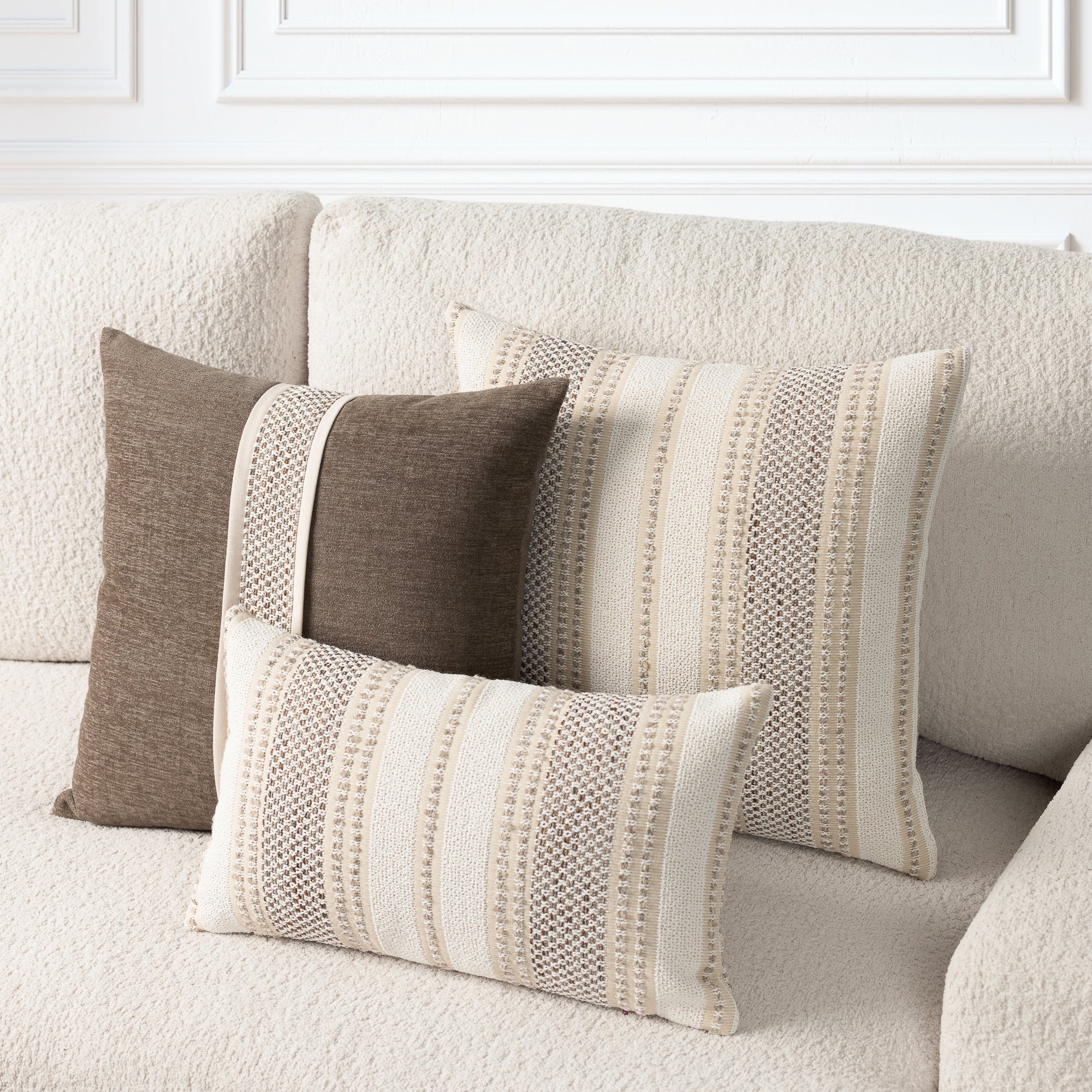 Three decorative throw pillows in neutral tones arranged on a textured cream sofa. Two larger pillows and one smaller lumbar pillow feature vertical stripe patterns in cream, beige, and taupe, with one solid taupe pillow on the left.