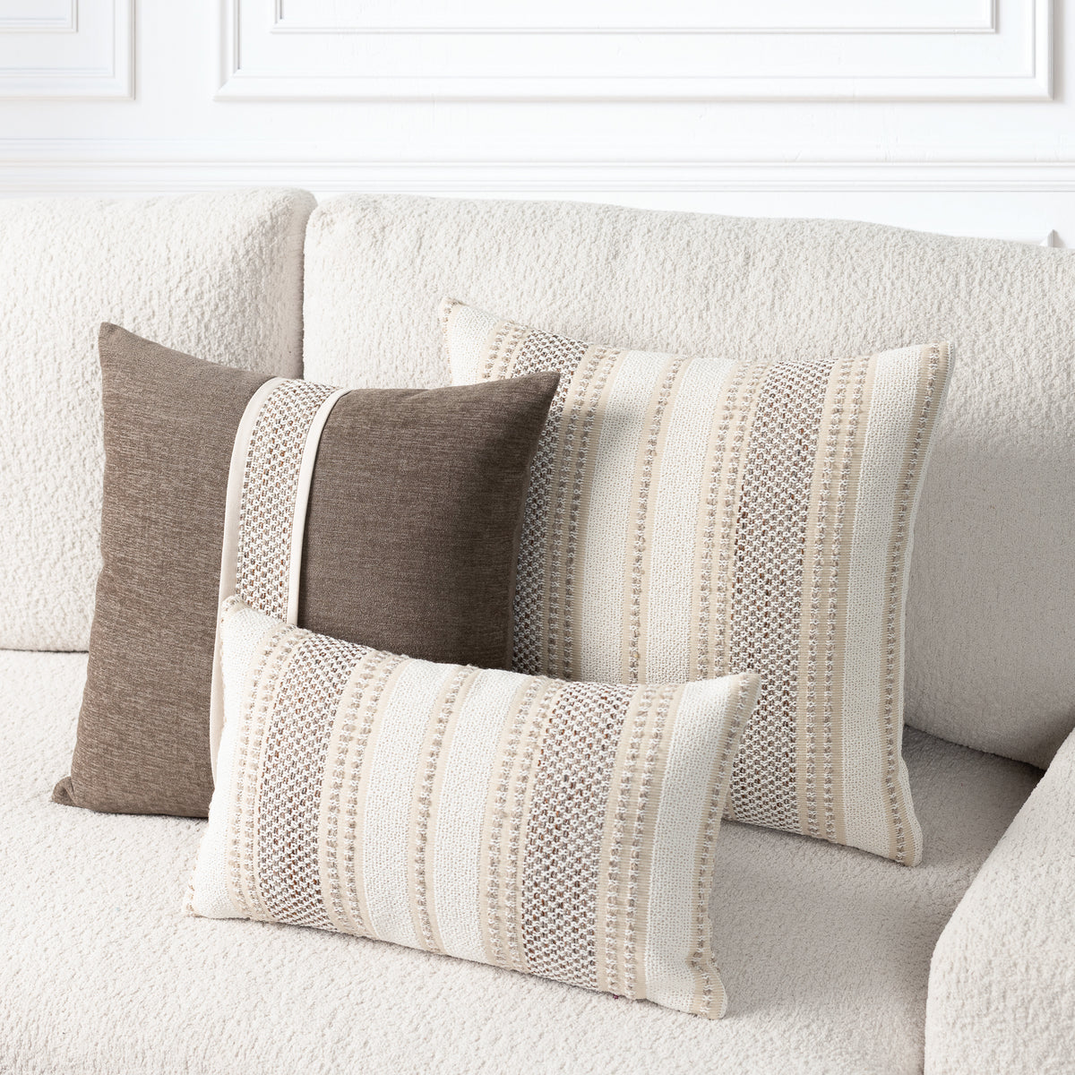 Three decorative throw pillows in neutral tones arranged on a textured cream sofa. Two larger pillows and one smaller lumbar pillow feature vertical stripe patterns in cream, beige, and taupe, with one solid taupe pillow on the left.
