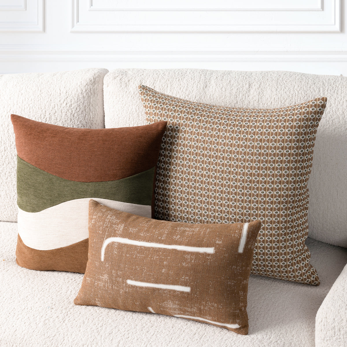 Three decorative throw pillows arranged on a cream textured sofa against a white paneled wall. The pillows include a rust brown and olive green color-blocked design, a small-scale geometric pattern in brown and beige, and a rust brown linen-style pillow with white abstract lines.