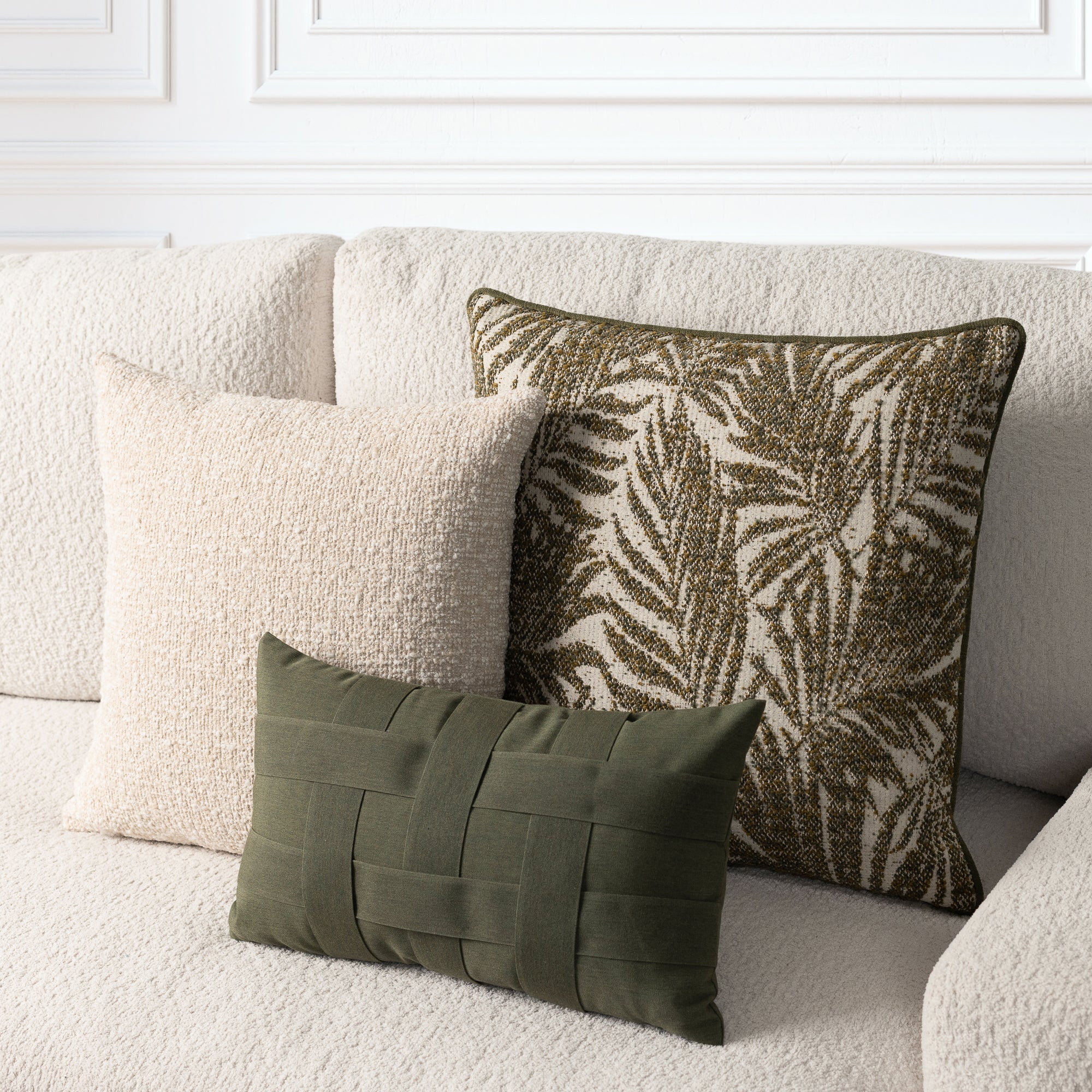 Three decorative throw pillows arranged on a textured cream boucle sofa: a large square pillow with an olive and cream palm leaf pattern, a smaller square cream pillow, and a small rectangular olive green woven lumbar pillow.