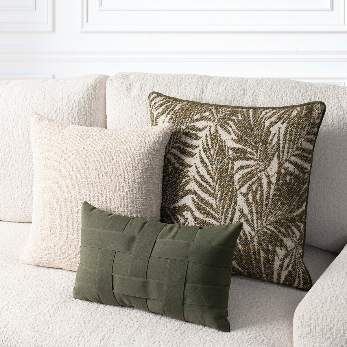 Three decorative throw pillows arranged on a textured cream boucle sofa: a large square pillow with an olive and cream palm leaf pattern, a smaller square cream pillow, and a small rectangular olive green woven lumbar pillow.
