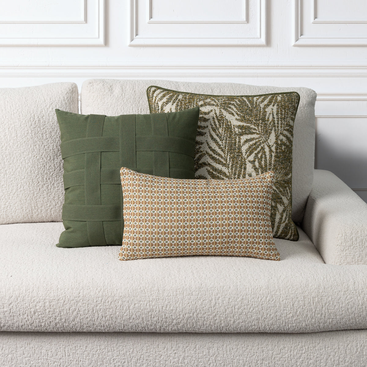 Three decorative throw pillows arranged on a light beige textured sofa against a white paneled wall. The pillows include an olive green woven square pillow, a green and beige palm leaf patterned square pillow, and a smaller rectangular pillow with a beige, gold, and green geometric pattern.