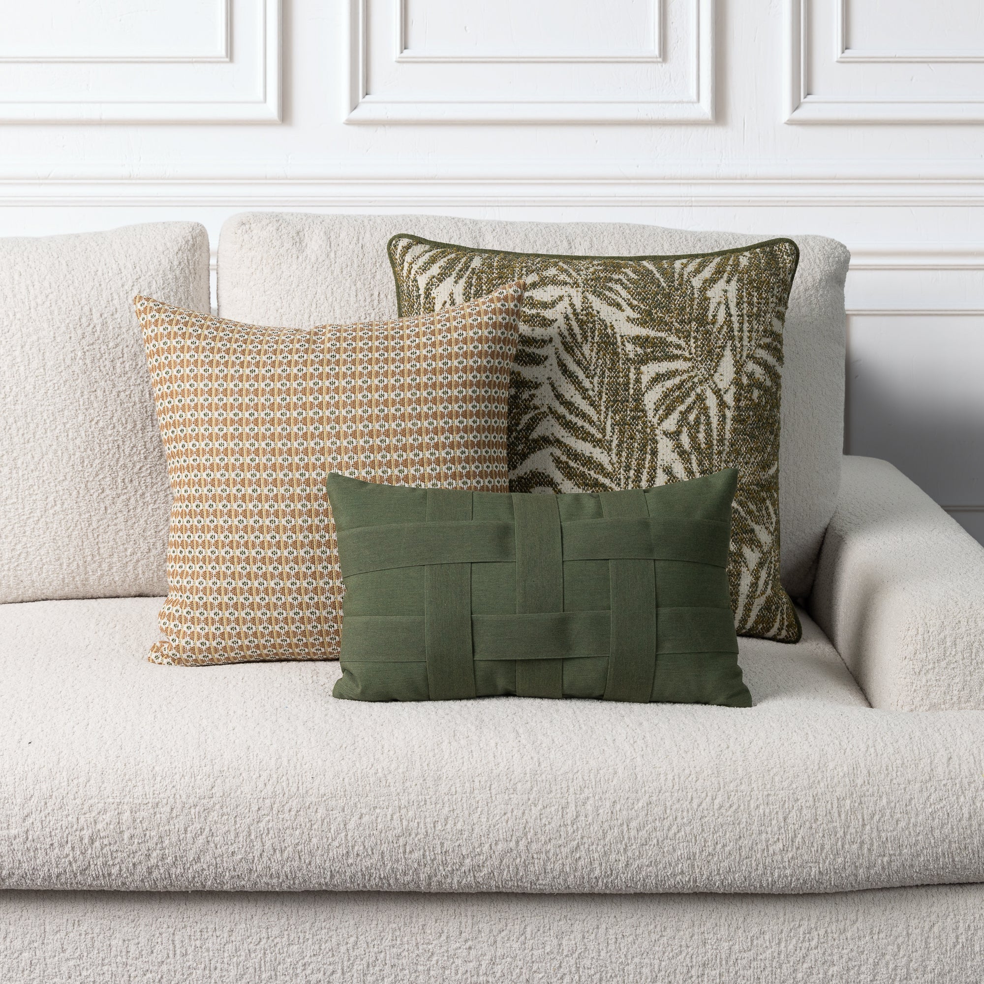 Three decorative throw pillows arranged on a light beige boucle sofa against a white paneled wall. The pillows include a green woven lumbar pillow, a tan and gold geometric pattern pillow, and an olive green palm leaf pattern pillow.