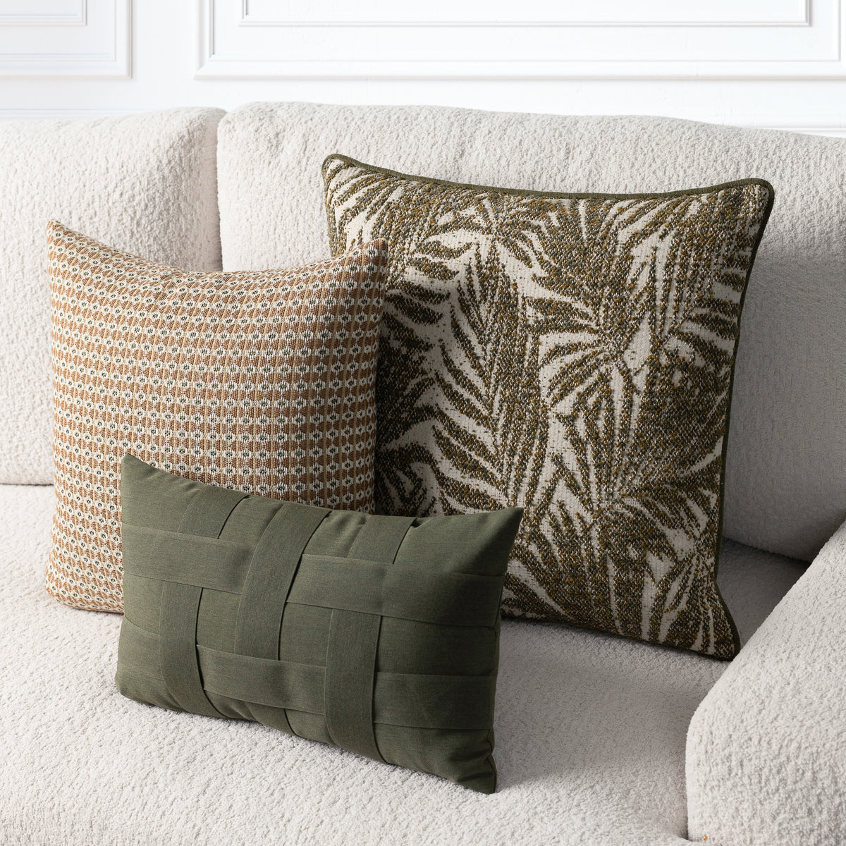 Three decorative throw pillows arranged on a textured cream sofa: a square olive green palm leaf pattern pillow, a rectangular beige and brown geometric pattern pillow, and a smaller square olive green woven basketweave pillow.