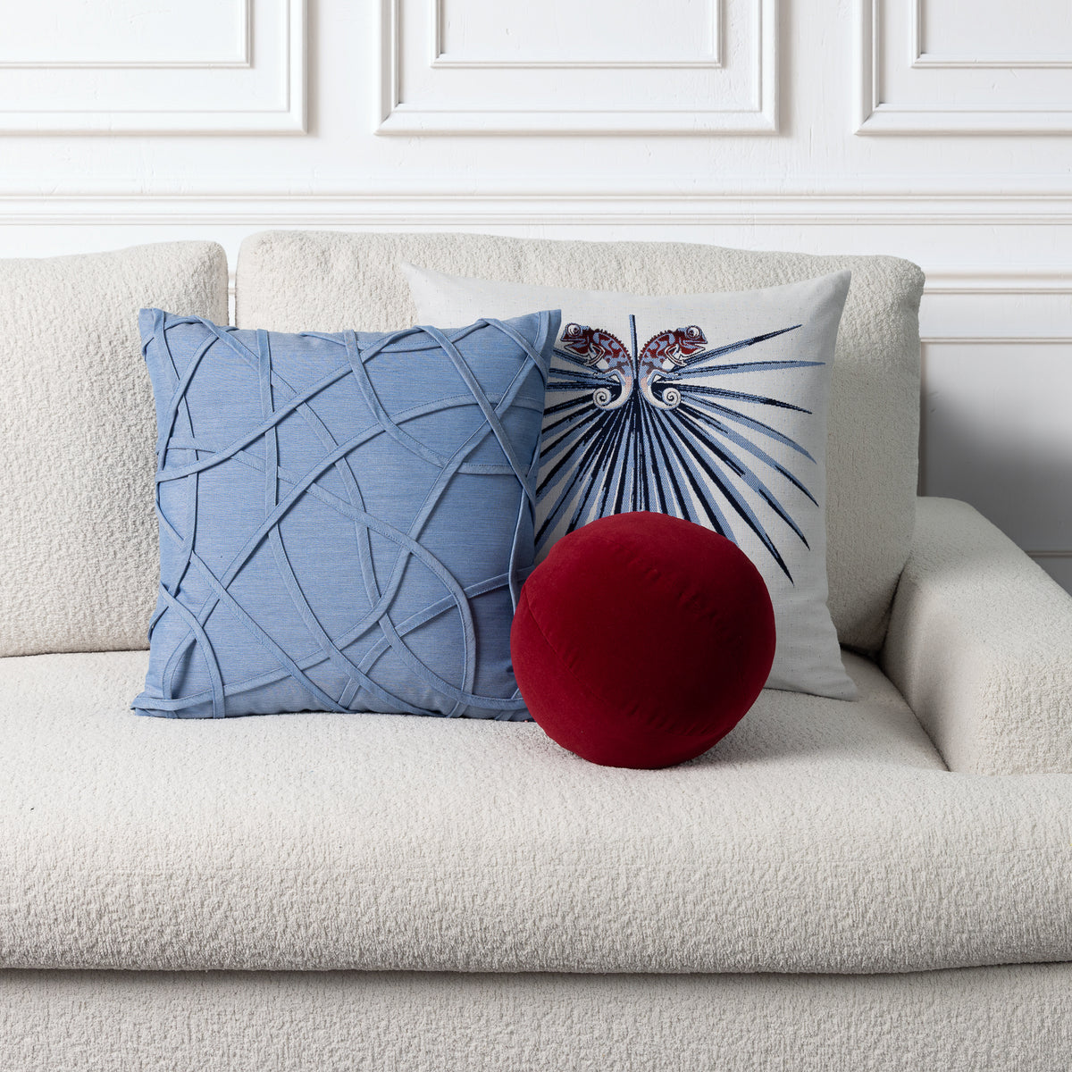 Three decorative throw pillows arranged on a textured cream sofa against a white paneled wall: a square light blue pillow with an abstract crisscross pattern, a square white pillow with a blue palm leaf design and butterfly motif, and a round deep red velvet pillow in front.