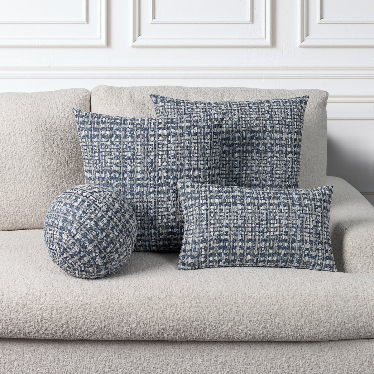 Three decorative throw pillows in a blue and white textured basketweave pattern arranged on a light beige boucle sofa against a white paneled wall.
