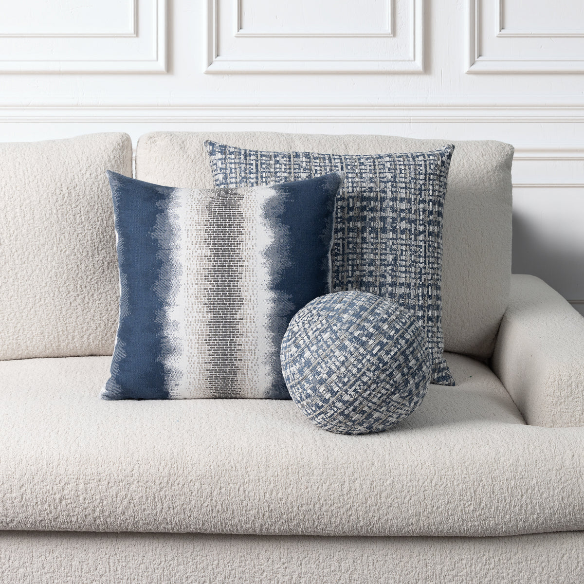 Three decorative throw pillows in blue, navy, and white textured fabrics arranged on a light beige boucle sofa against a white paneled wall.