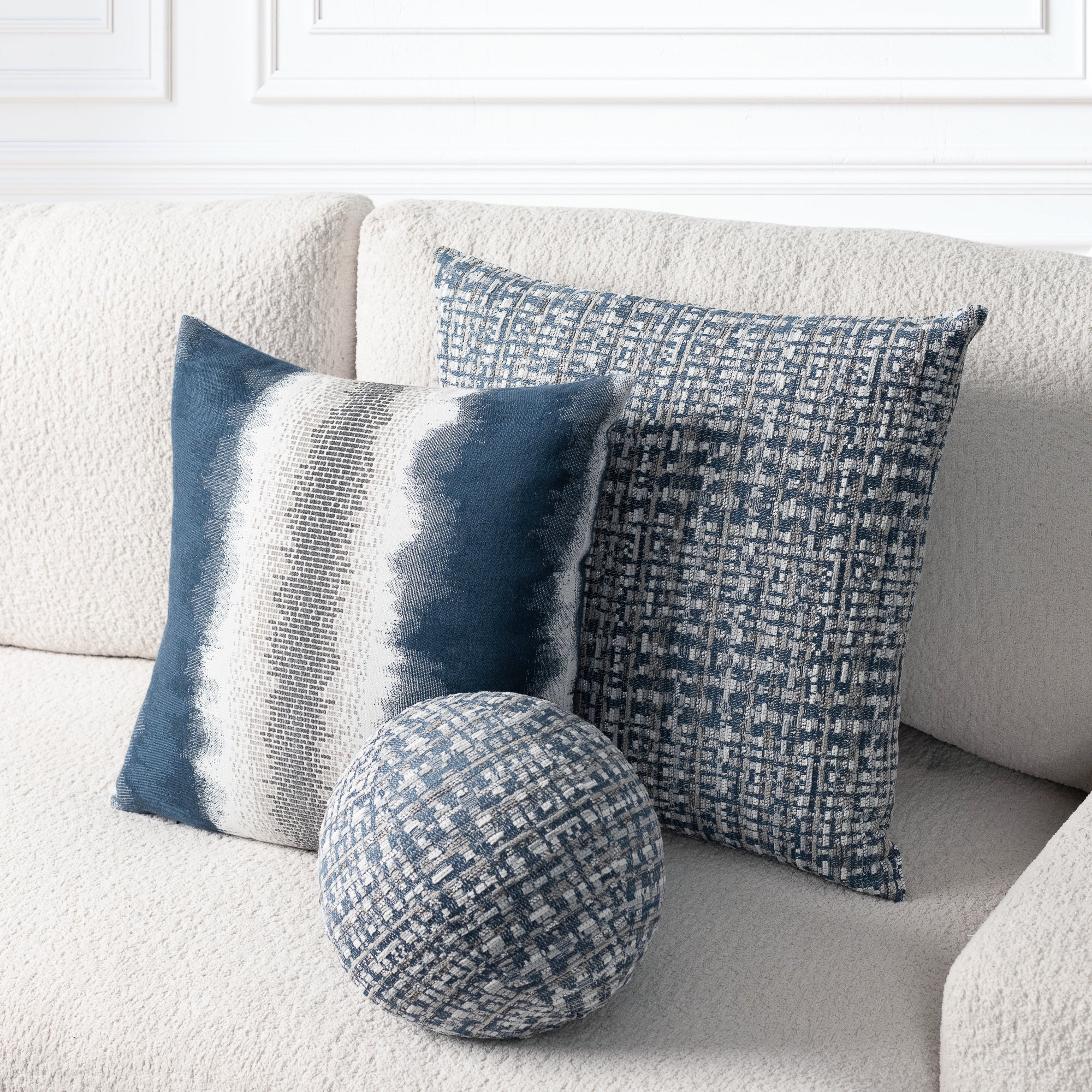 Three decorative throw pillows in blue, navy, and gray tones arranged on a textured cream sofa. The set includes two square pillows with woven patterns and one round matching accent pillow.