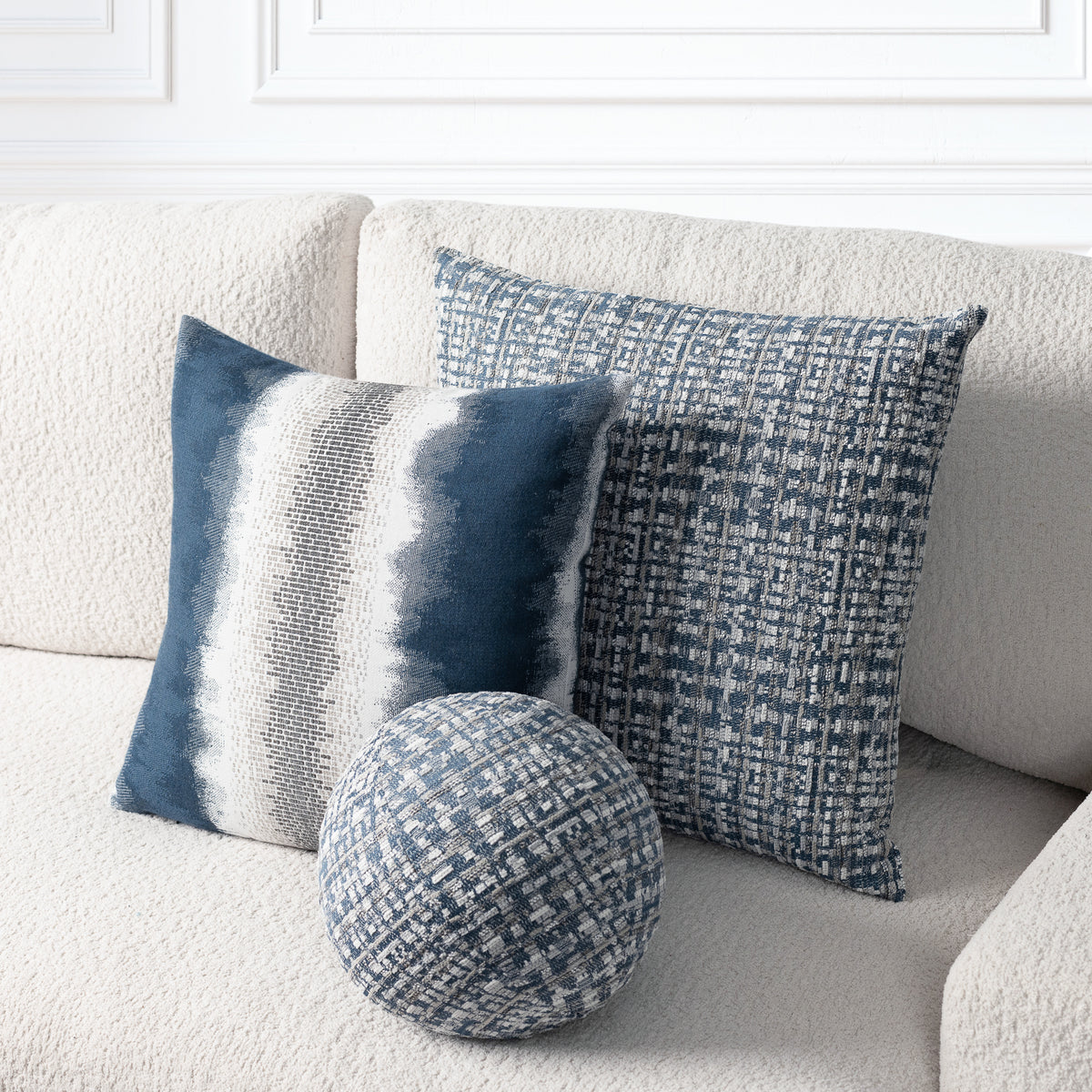 Three decorative throw pillows in blue, navy, and gray tones arranged on a textured cream sofa. The set includes two square pillows with woven patterns and one round matching accent pillow.