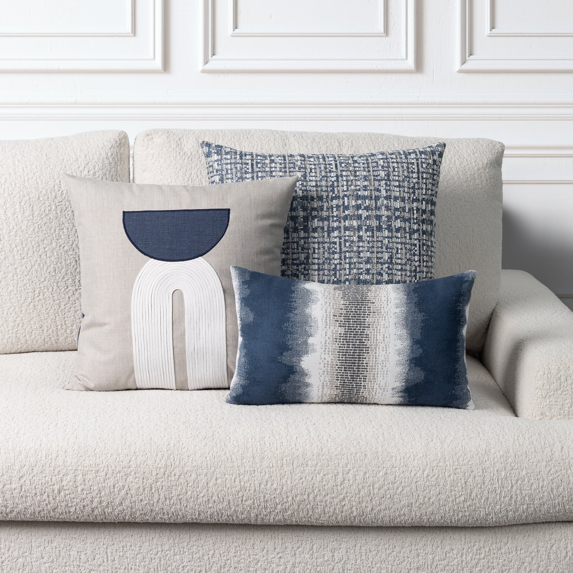 Three decorative throw pillows arranged on a textured cream boucle sofa against a white paneled wall. The pillows include a large beige pillow with a navy and white abstract design, a blue-and-white woven pattern pillow, and a blue-and-white striped lumbar pillow.