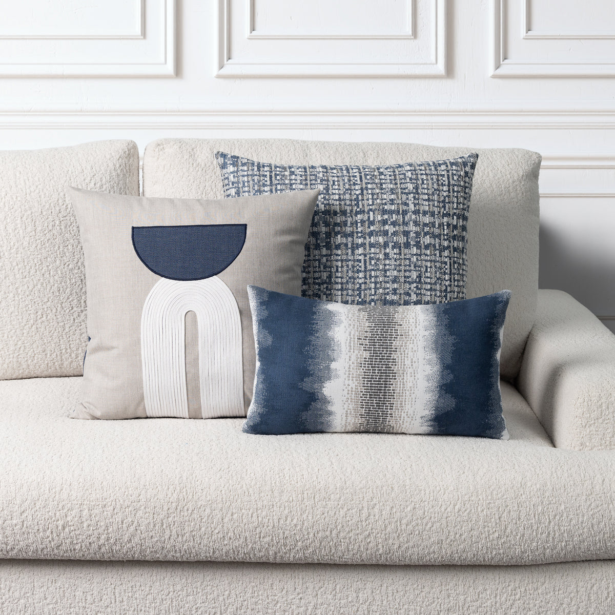 Three decorative throw pillows arranged on a textured cream boucle sofa against a white paneled wall. The pillows include a large beige pillow with a navy and white abstract design, a blue-and-white woven pattern pillow, and a blue-and-white striped lumbar pillow.