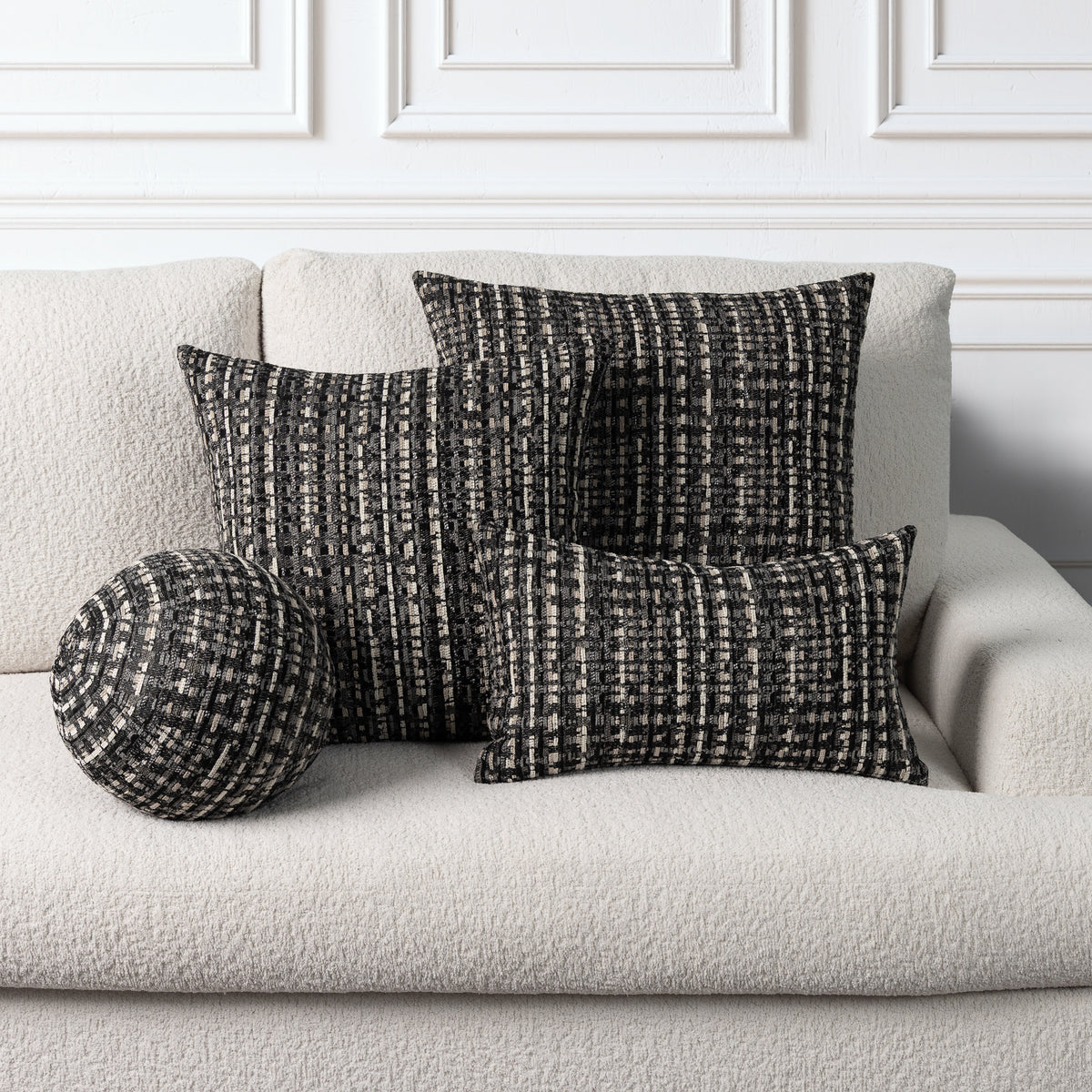 Three textured throw pillows in a black and off-white woven pattern arranged on a light beige boucle sofa against a white paneled wall.
