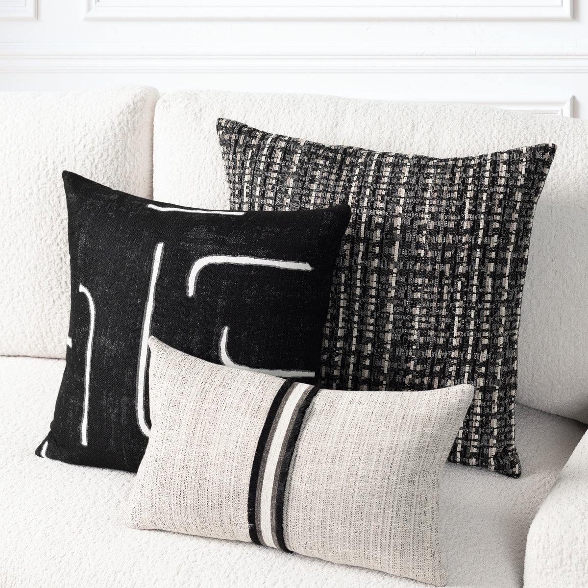 Three decorative throw pillows arranged on a textured off-white sofa. The pillows include a black square with white abstract lines, a black and gray woven square, and a light beige rectangular pillow with two black stripes.