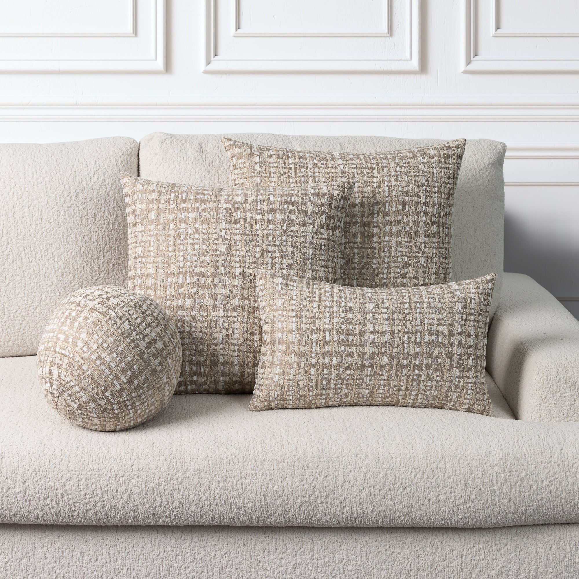 Three textured throw pillows in neutral beige and cream tones arranged on a light beige sofa against a white paneled wall.
