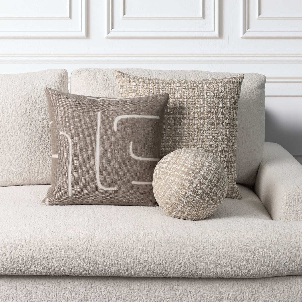 Three neutral-toned decorative throw pillows arranged on a light beige sofa against a white paneled wall. One square pillow has a taupe background with white abstract line art, one square pillow has a woven beige texture, and one round pillow has a textured cream-and-beige pattern.