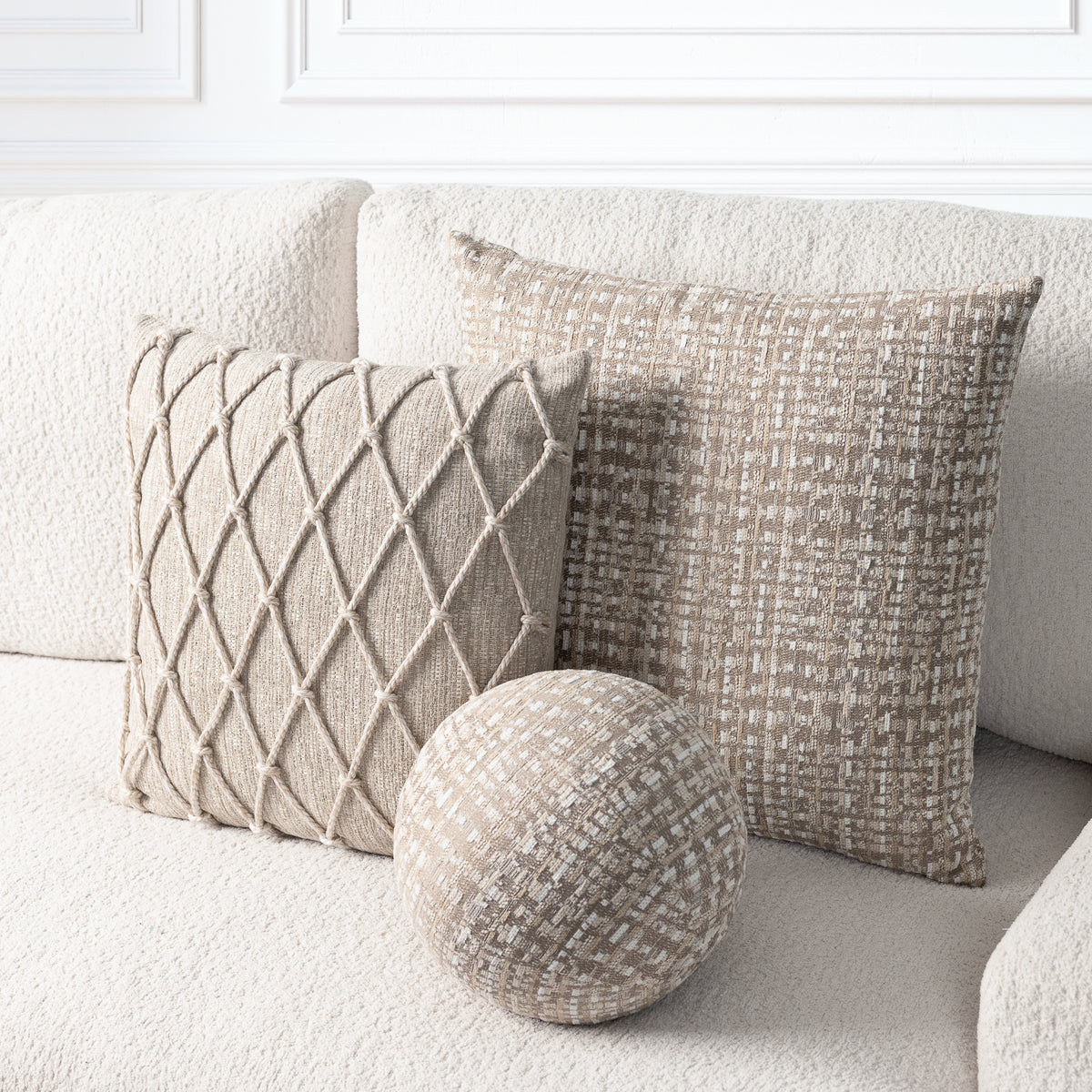 Three neutral-toned decorative pillows on a cream boucle sofa: a square woven throw, a textured square throw, and a round woven throw, styled in a bright living room setting.