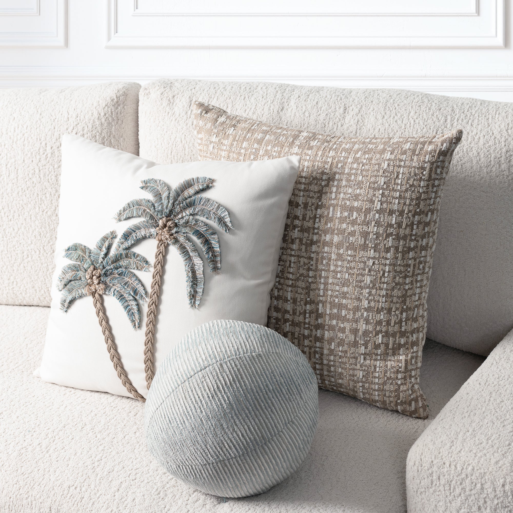 A styled home decor scene showing two decorative throw pillows and a round lumbar pillow on a textured beige sofa. The front pillow is off-white with a raised palm tree motif in blue-gray and tan.