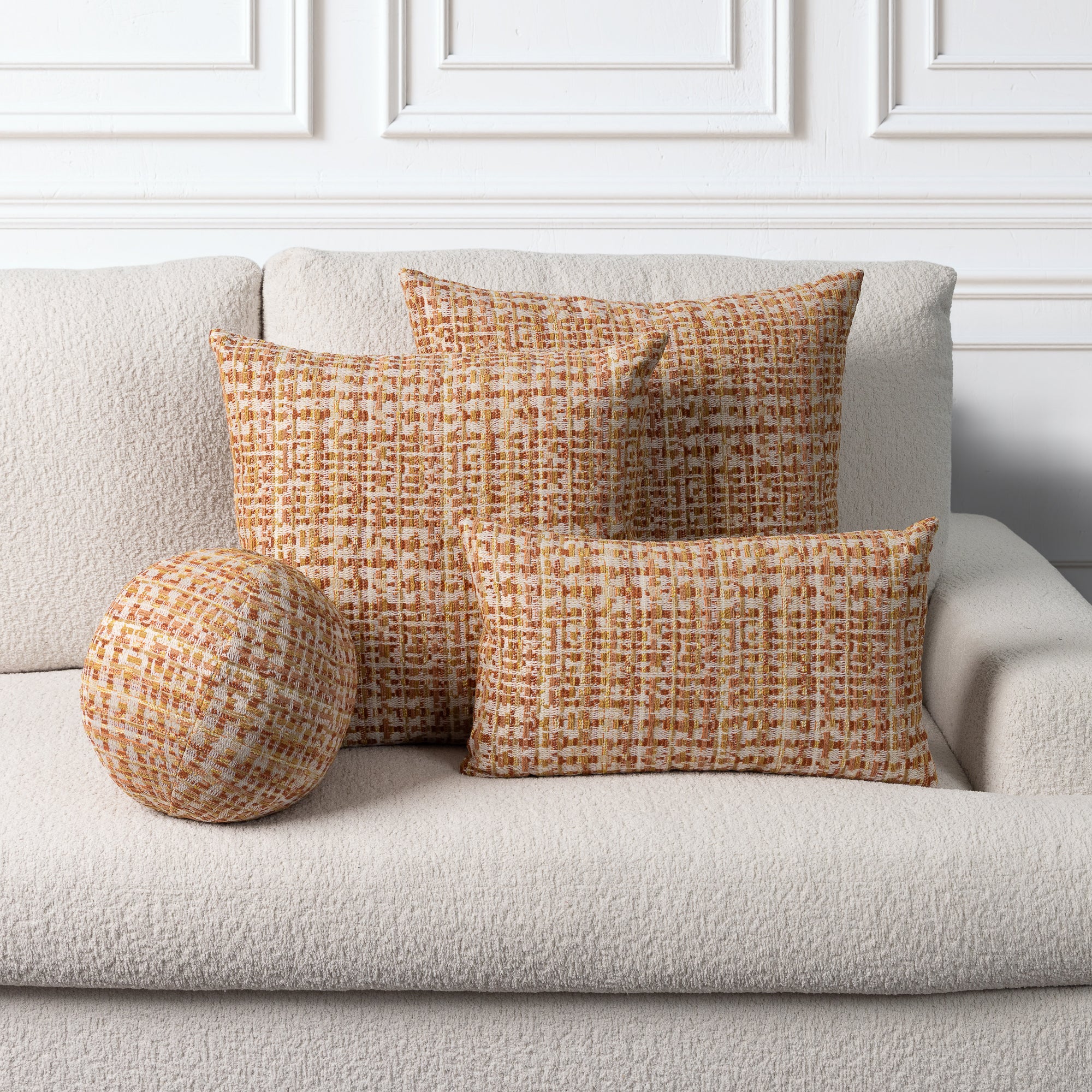 Three textured throw pillows in a warm orange and cream bouclé pattern arranged on a light beige sofa against a white paneled wall.