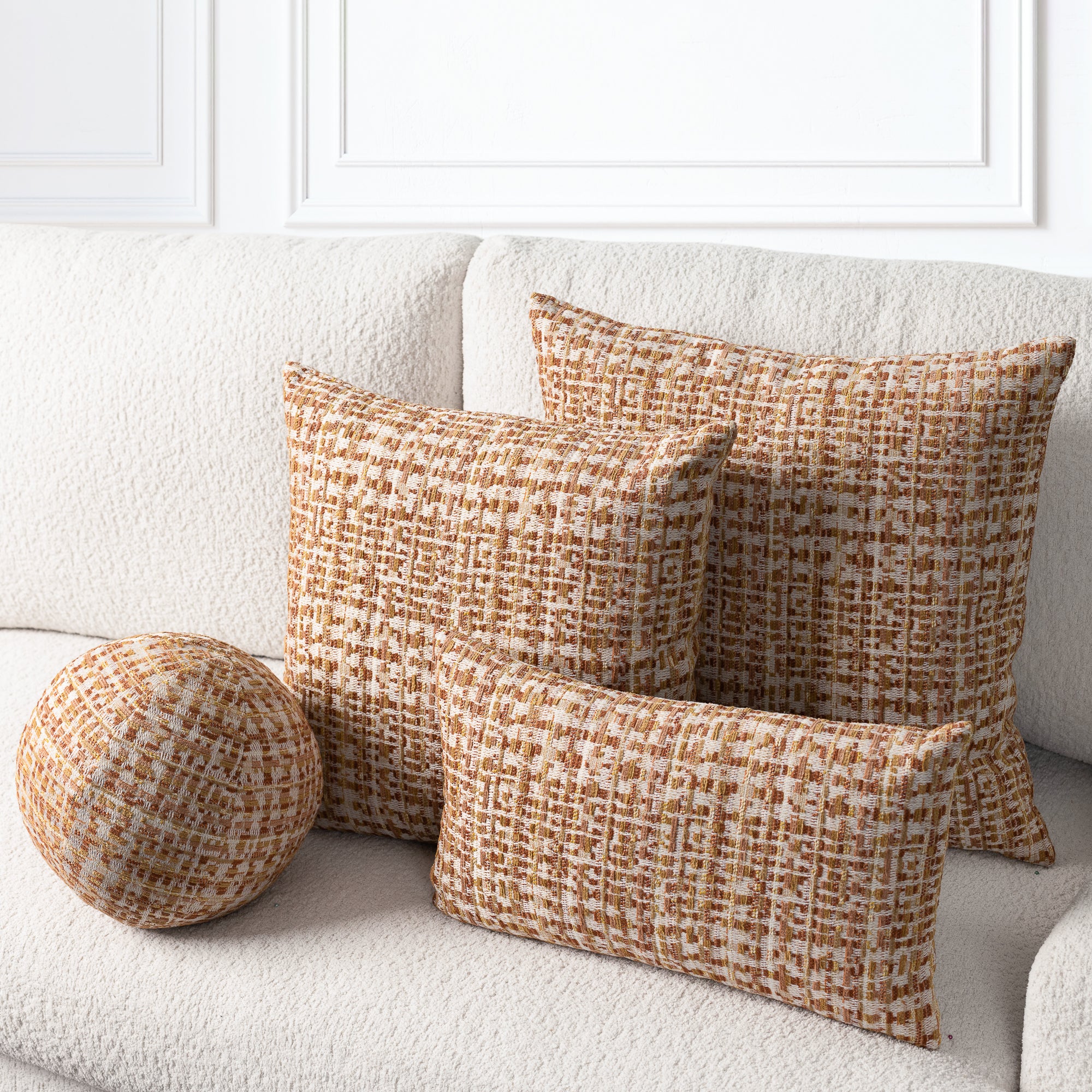 Three woven throw pillows in a warm brown and cream basketweave pattern arranged on a textured off-white sofa, including two square pillows, one rectangular lumbar pillow, and one round bolster pillow.