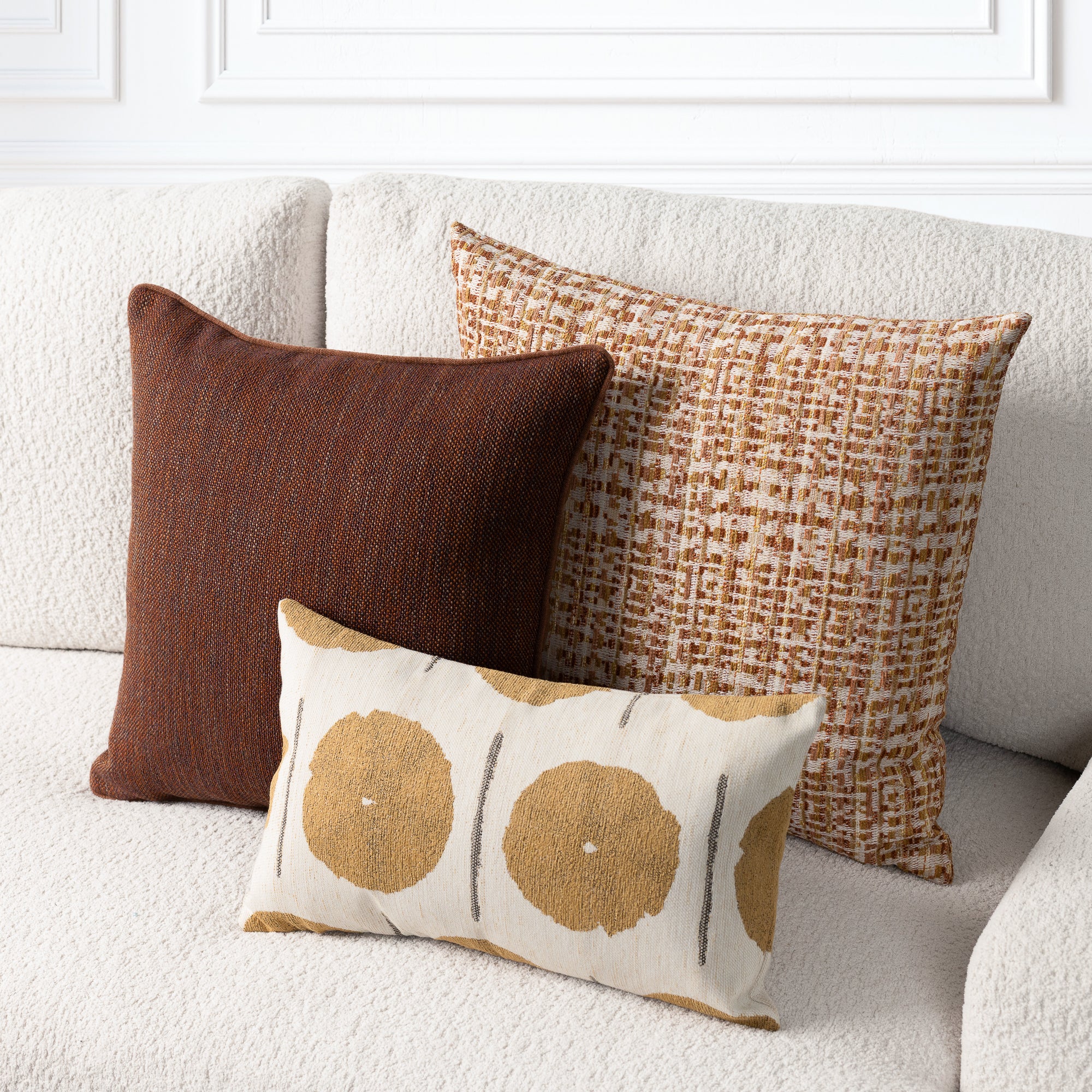 Three decorative throw pillows arranged on a textured cream sofa: a square dark brown pillow, a square tan and brown woven pattern pillow, and a smaller rectangular cream pillow with large tan circular motifs and thin gray vertical lines.
