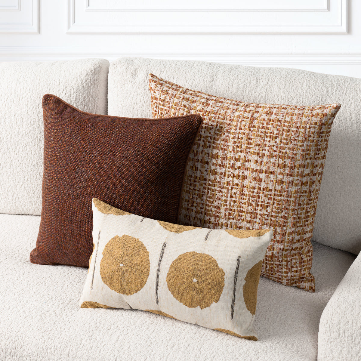 Three decorative throw pillows arranged on a textured cream sofa: a square dark brown pillow, a square tan and brown woven pattern pillow, and a smaller rectangular cream pillow with large tan circular motifs and thin gray vertical lines.