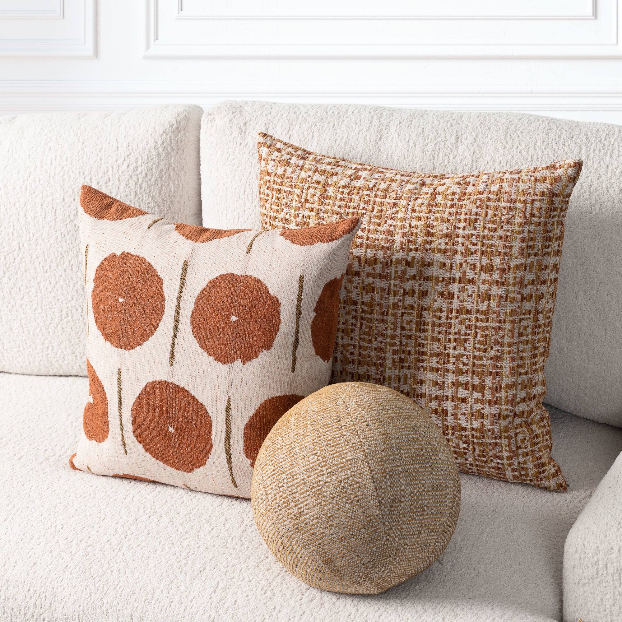 Three decorative throw pillows arranged on a cream boucle sofa: a square pillow with large rust-brown circular motifs on a light beige background, a textured square pillow in warm tan and brown, and a round woven jute pillow in front.