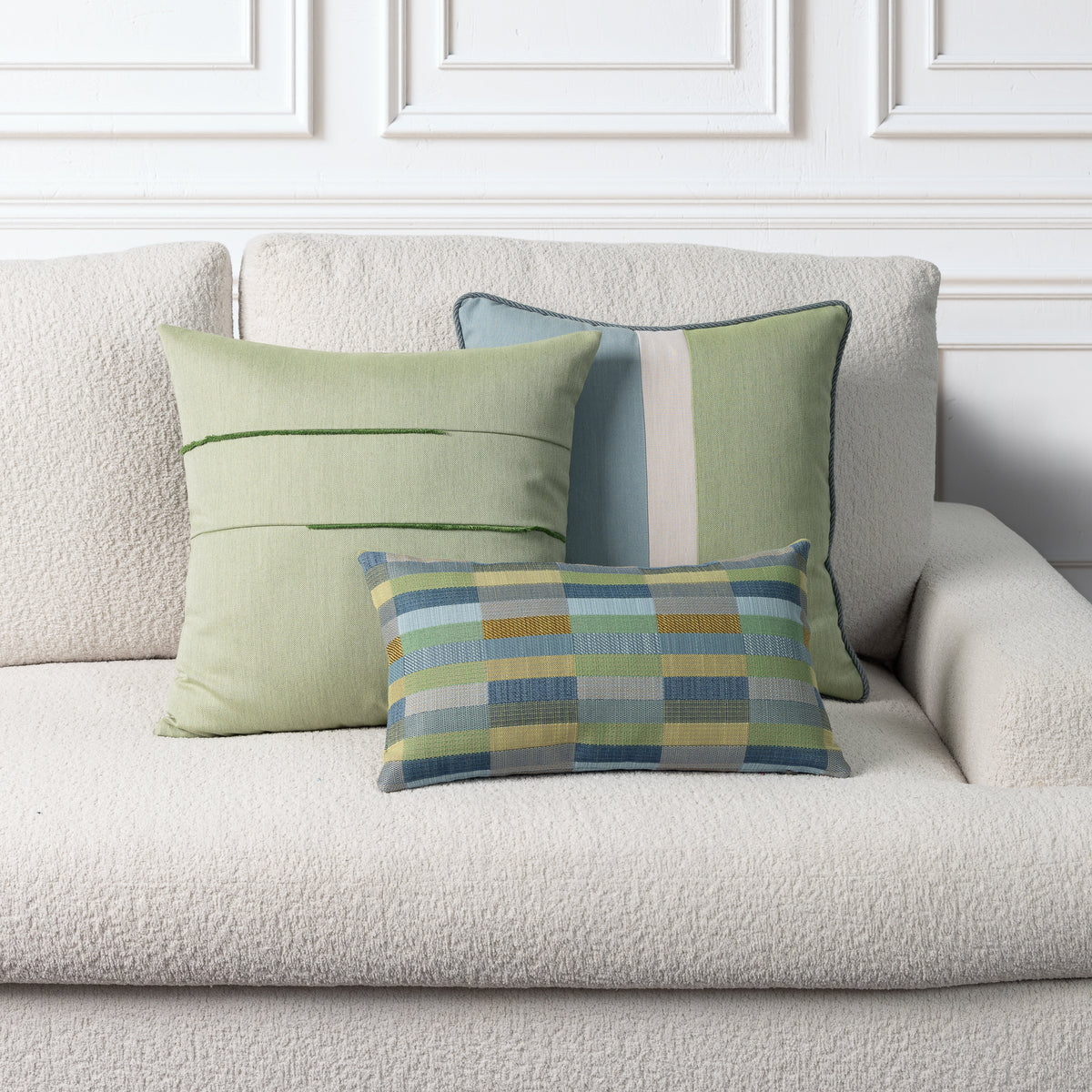 Three decorative throw pillows in shades of sage green, blue, and yellow are arranged on a textured cream boucle sofa against a white paneled wall.