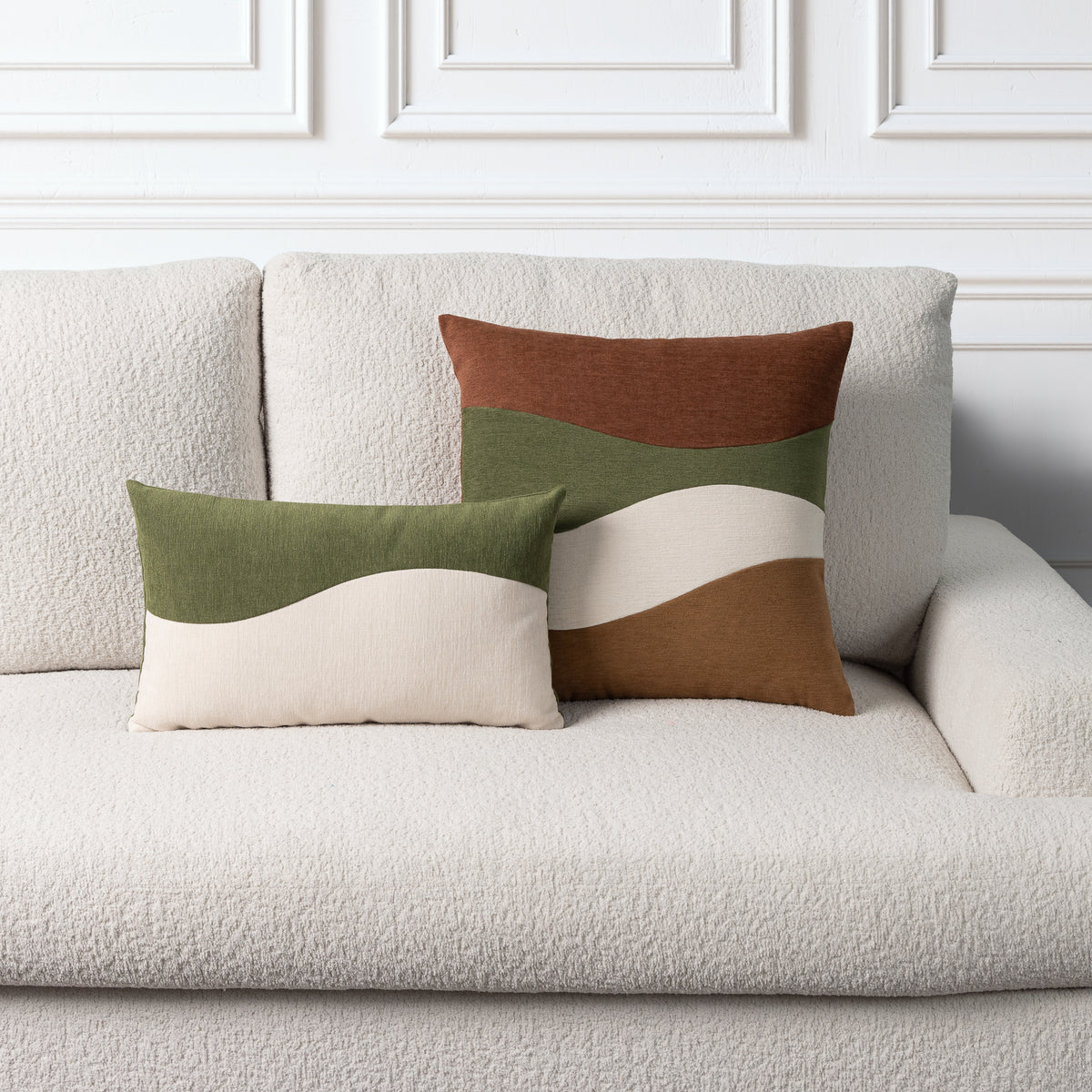 Two decorative throw pillows with wavy color-blocked stripes in olive green, cream, and brown, styled on a textured off-white boucle sofa against a white paneled wall.