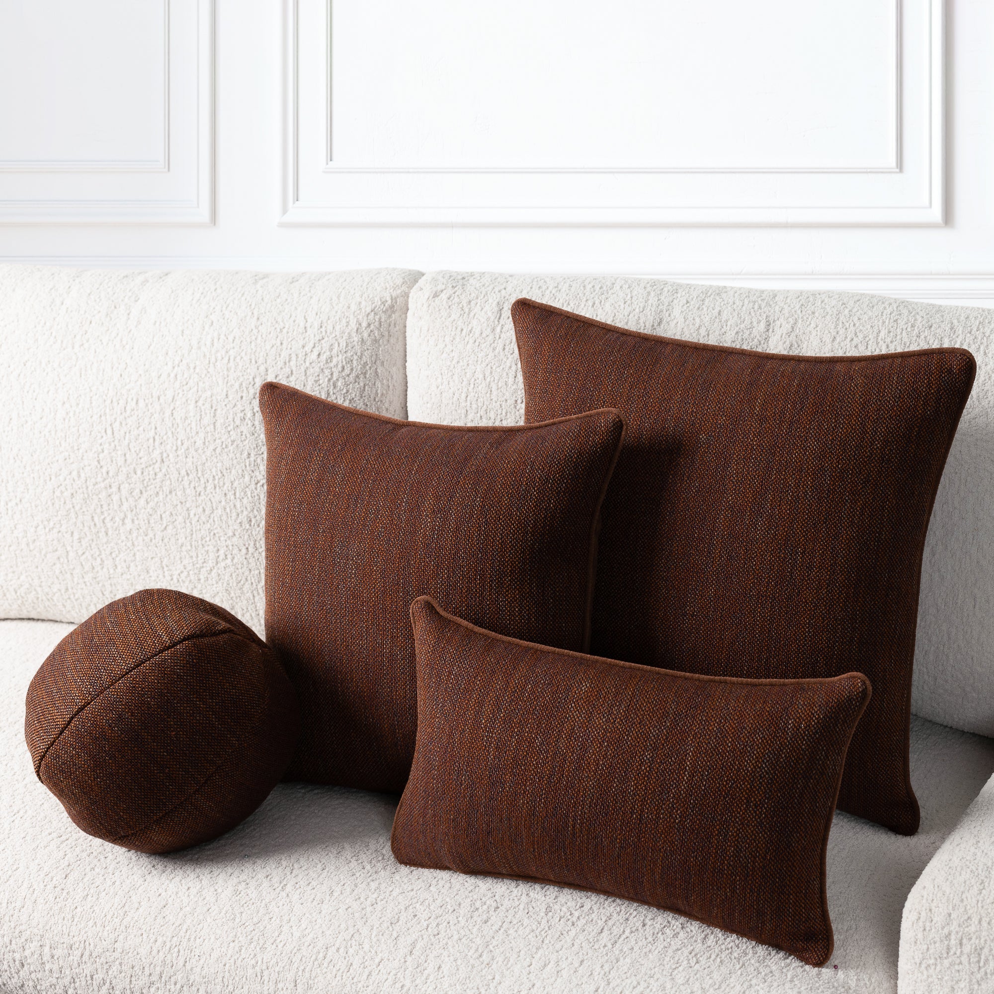 Four dark brown textured throw pillows, including three square pillows and one round lumbar pillow, arranged on a white boucle sofa against a white paneled wall.