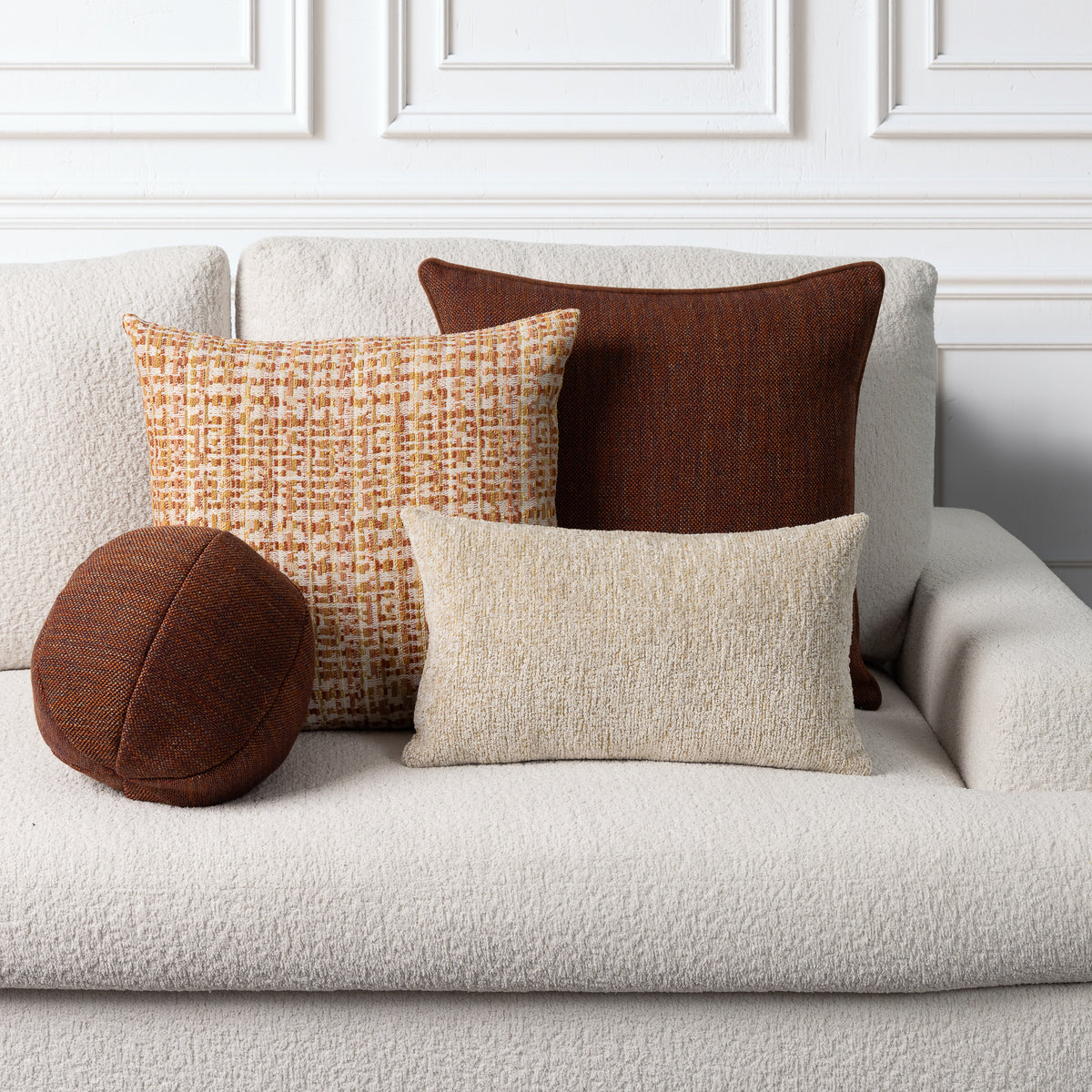 Four decorative throw pillows in neutral and warm tones arranged on a light beige boucle sofa against a white paneled wall.