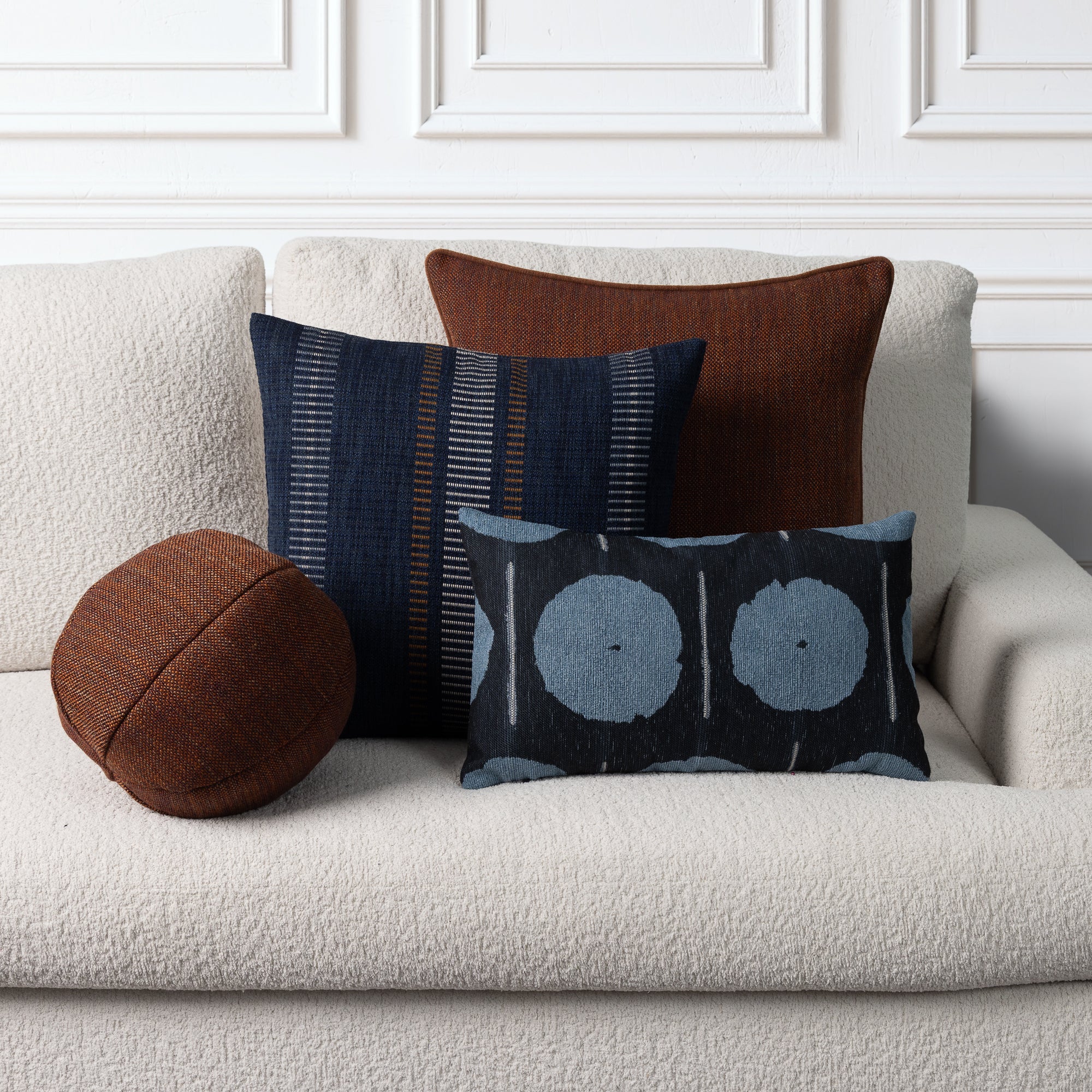 A styled collection of home decor throw pillows arranged on a textured cream boucle sofa against a white paneled wall. The set includes a round brown pillow, a dark blue striped pillow, a solid brown pillow, and a dark blue rectangular pillow with a light blue circular pattern.
