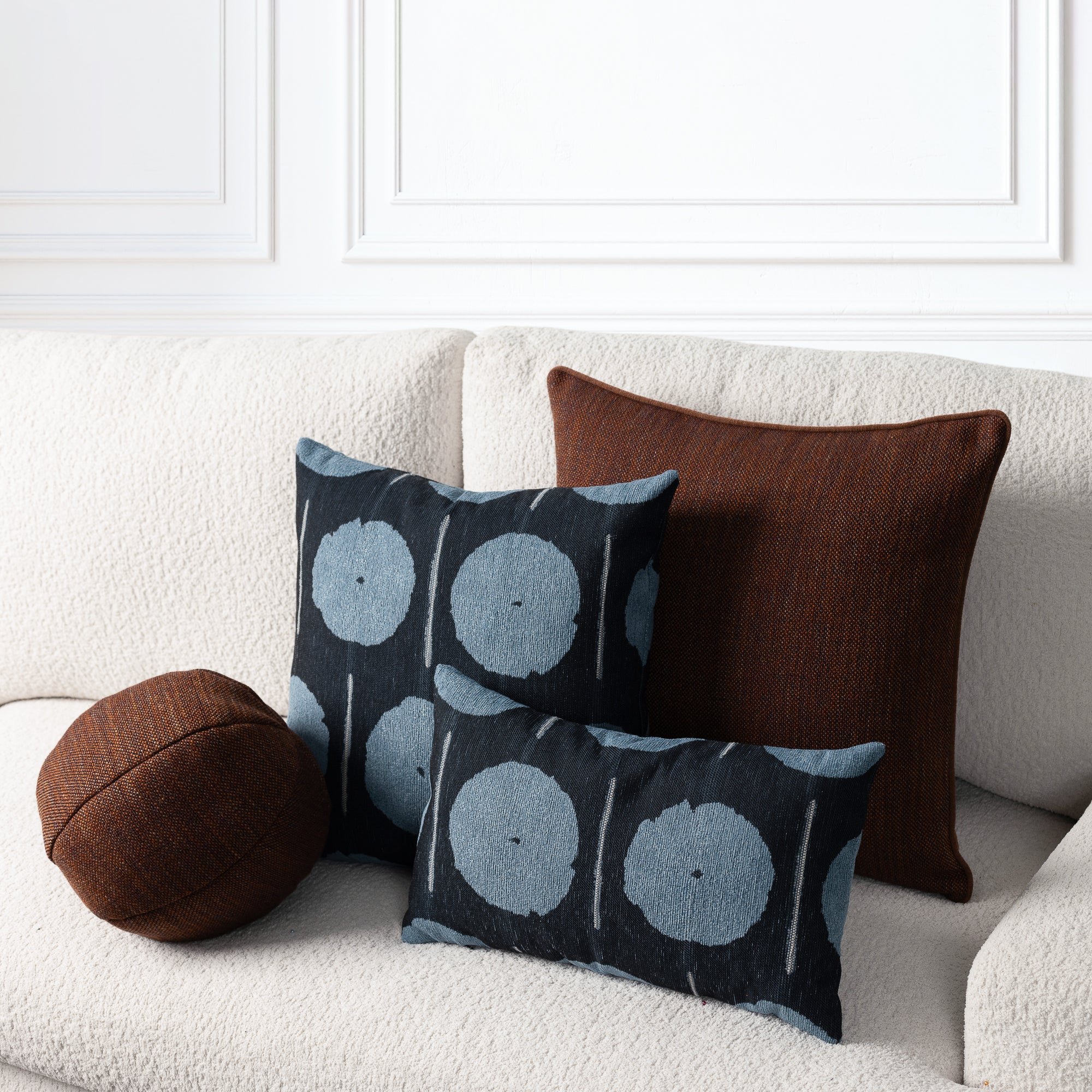 Three decorative throw pillows arranged on a cream boucle sofa against a white paneled wall. The set includes a round brown pillow, a square dark brown pillow, and two black pillows with a blue-gray circular ikat pattern.