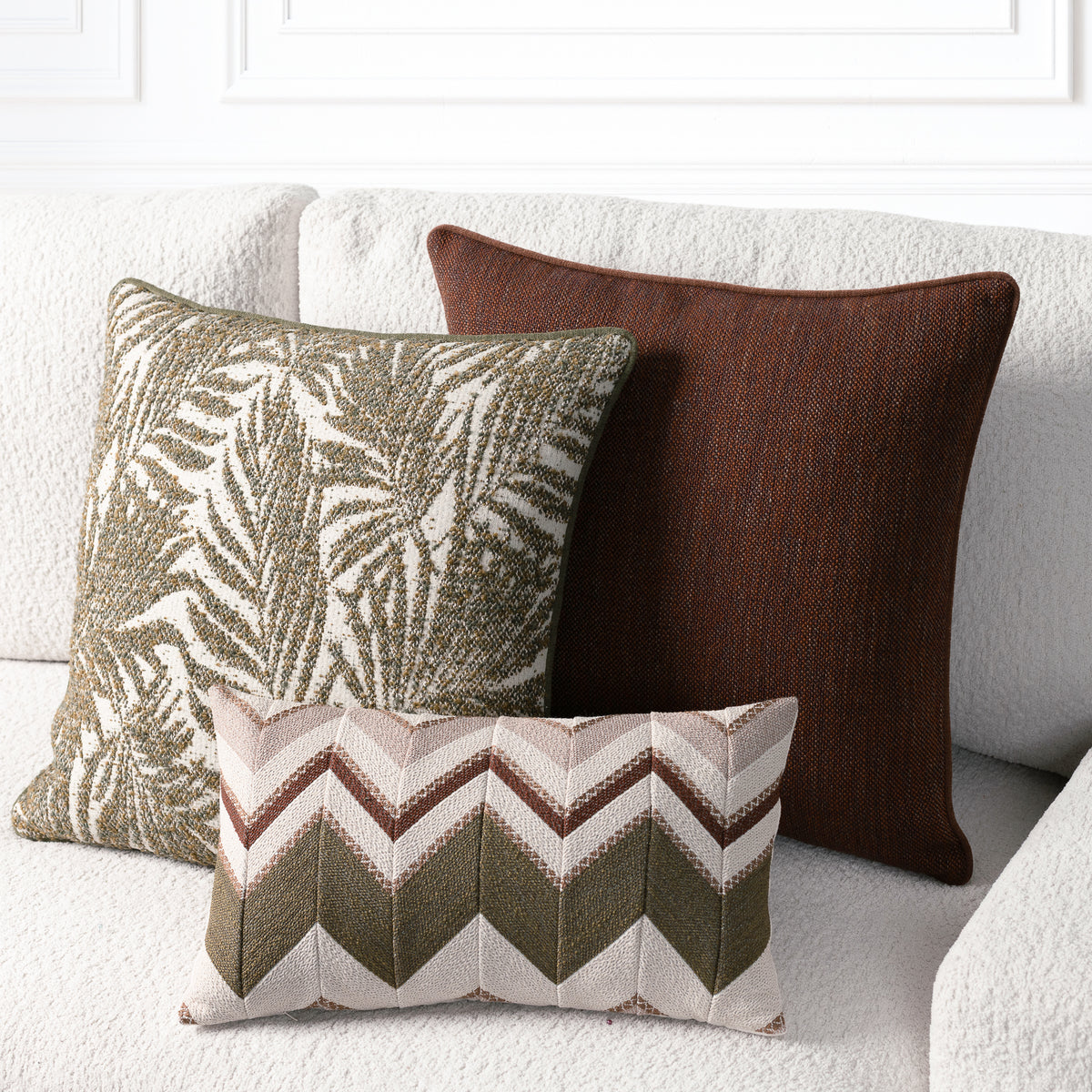 Three decorative throw pillows arranged on a light textured sofa: a large dark brown pillow, a green-and-cream palm leaf patterned pillow, and a small rectangular chevron pillow in beige, brown, and green.