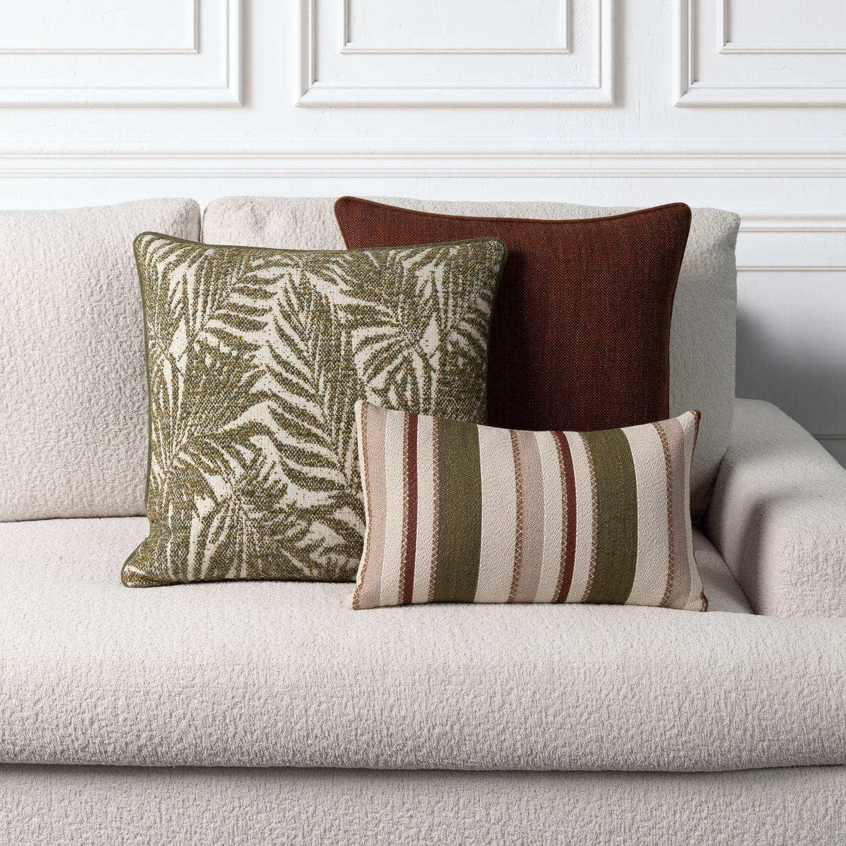 Three decorative throw pillows arranged on a light beige textured sofa against a white paneled wall. The pillows include a square olive green and cream palm leaf patterned pillow, a dark brown square pillow, and a smaller rectangular striped pillow in beige, olive green, and brown.