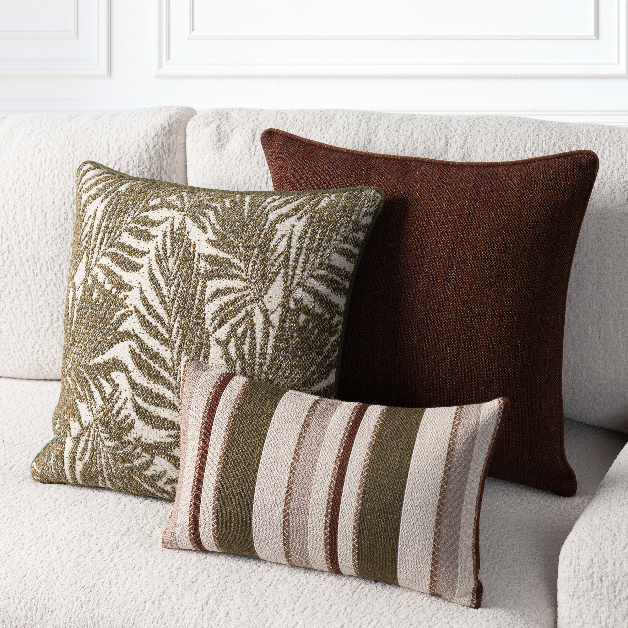 Three decorative throw pillows arranged on a light beige sofa: a large dark brown textured pillow, a square olive green and cream palm leaf patterned pillow, and a smaller rectangular striped pillow in olive green, cream, and brown.