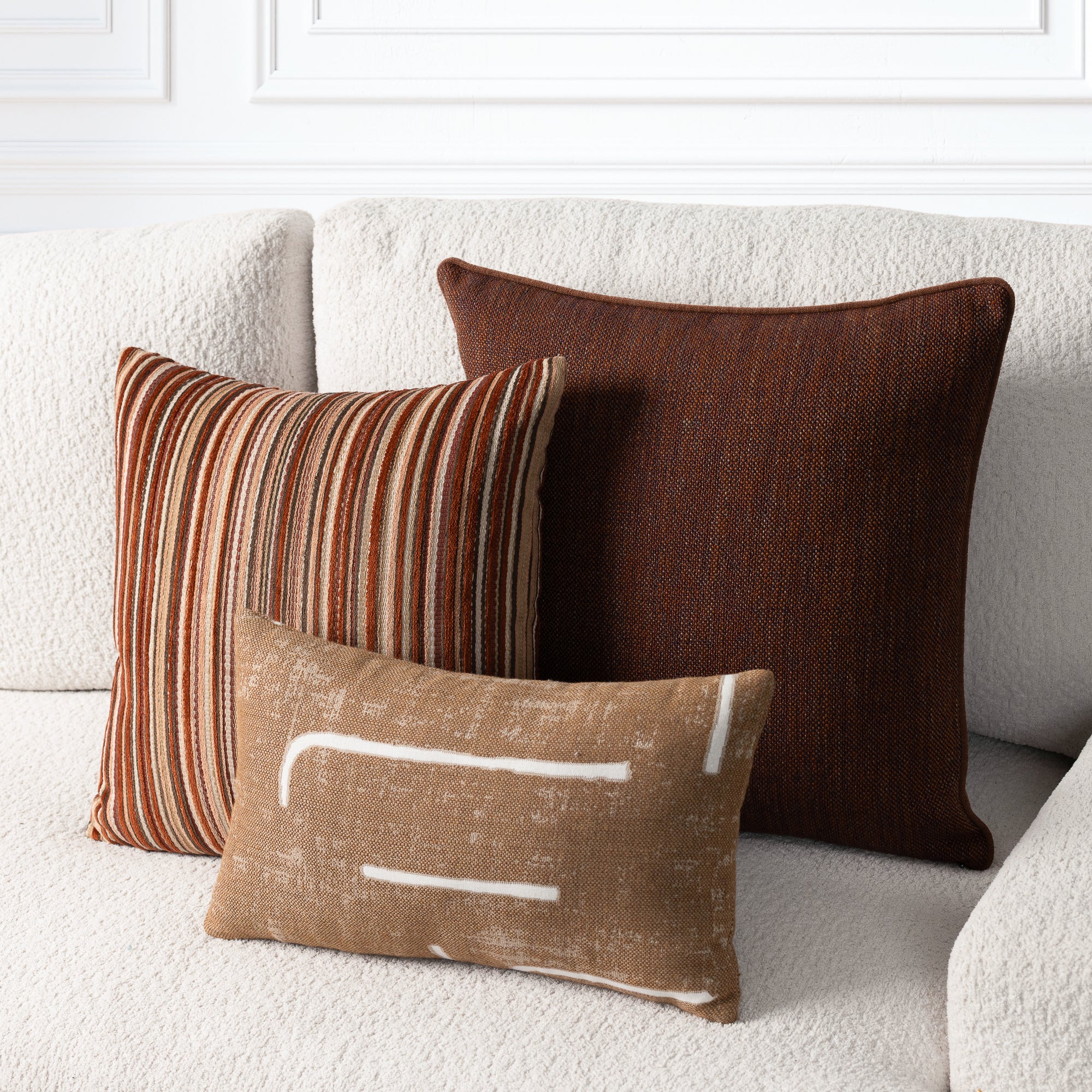 Three decorative throw pillows arranged on a textured cream boucle sofa: a large dark brown square pillow, a smaller striped rust, tan, and brown pillow, and a small tan lumbar pillow with white abstract lines.