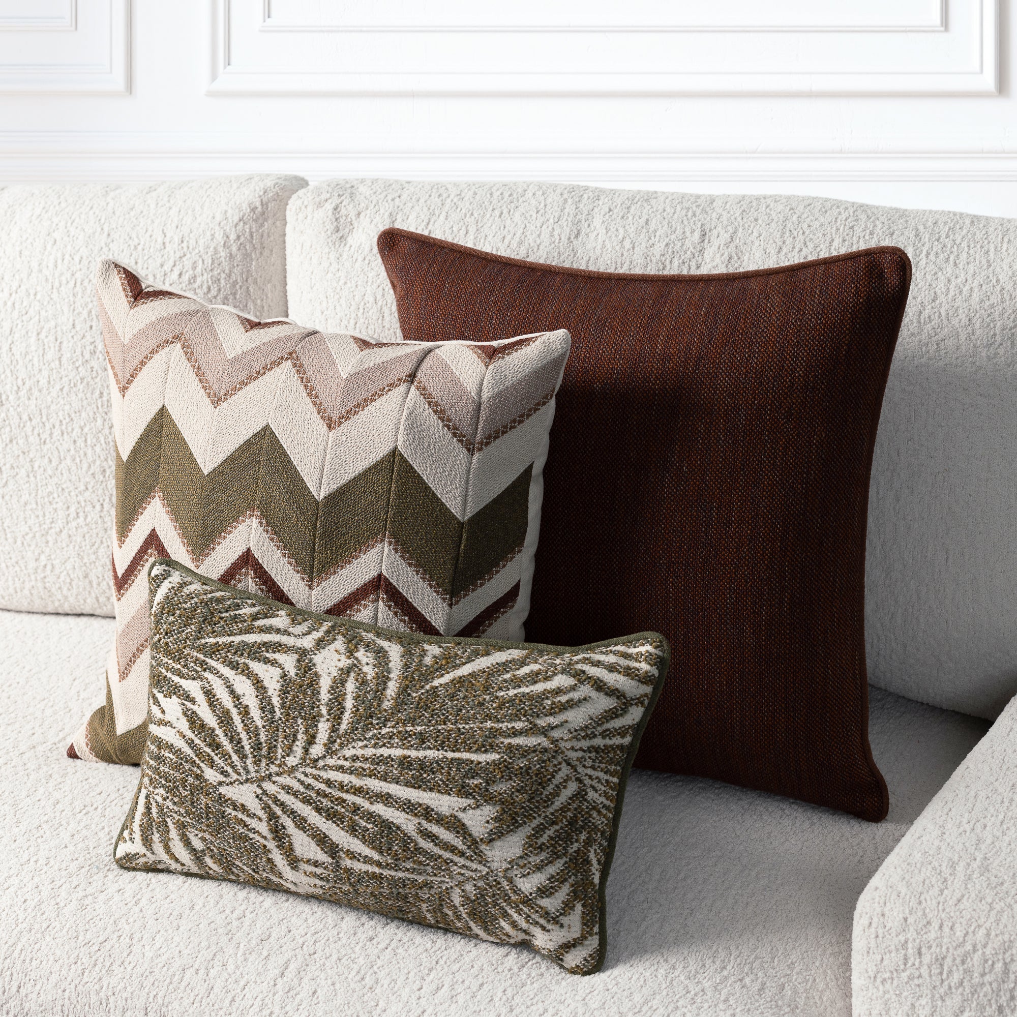 Three decorative throw pillows arranged on a light beige textured sofa: a large dark brown pillow, a square chevron-patterned pillow in beige, brown, and olive green, and a smaller rectangular pillow with a brown and beige leaf pattern.