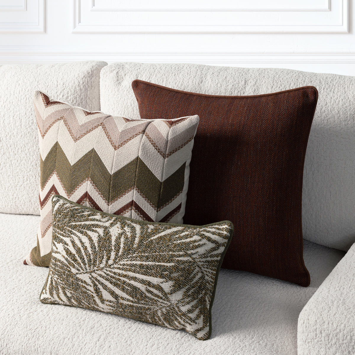Three decorative throw pillows arranged on a light beige textured sofa: a large dark brown pillow, a square chevron-patterned pillow in beige, brown, and olive green, and a smaller rectangular pillow with a brown and beige leaf pattern.