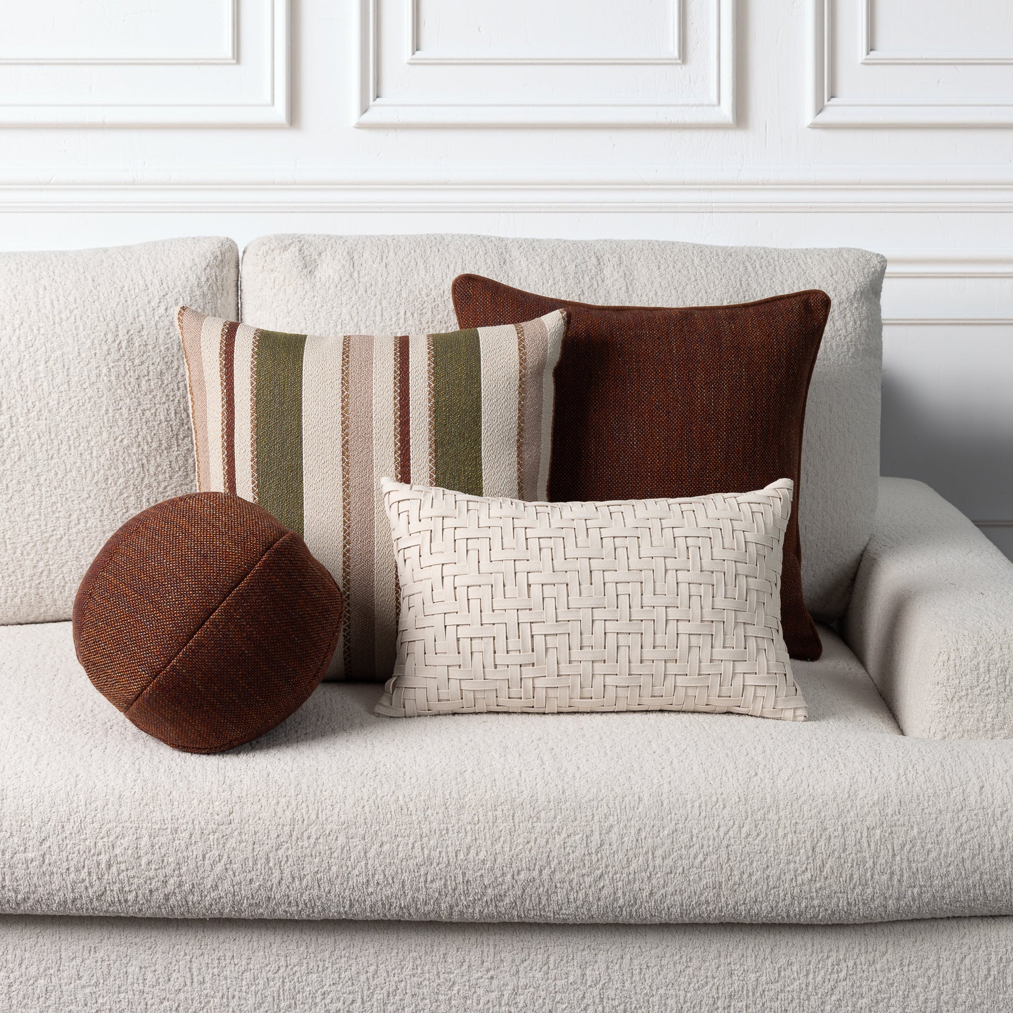 A set of five decorative throw pillows in various shapes and textures arranged on a textured cream boucle sofa against a white paneled wall.