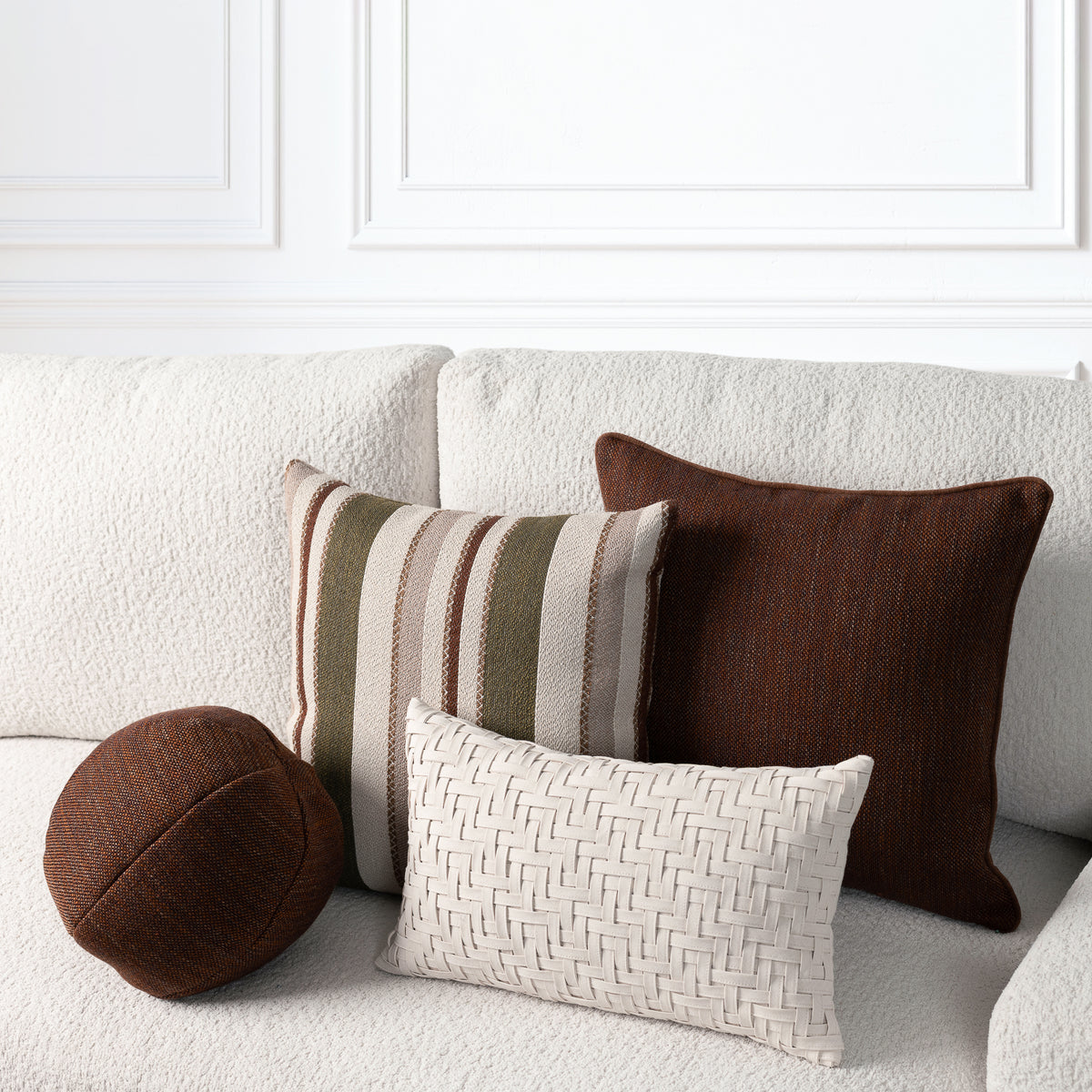 Four decorative throw pillows arranged on a cream boucle sofa against a white paneled wall. The set includes a round brown pillow, a square striped pillow in beige, brown, and olive green, a solid dark brown pillow, and a small square cream woven basket-texture pillow.