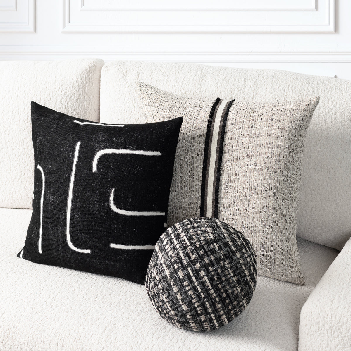 Three decorative throw pillows arranged on a textured white sofa: a black square pillow with white abstract line art, a light beige linen pillow with two vertical black stripes, and a round woven black-and-beige accent pillow.