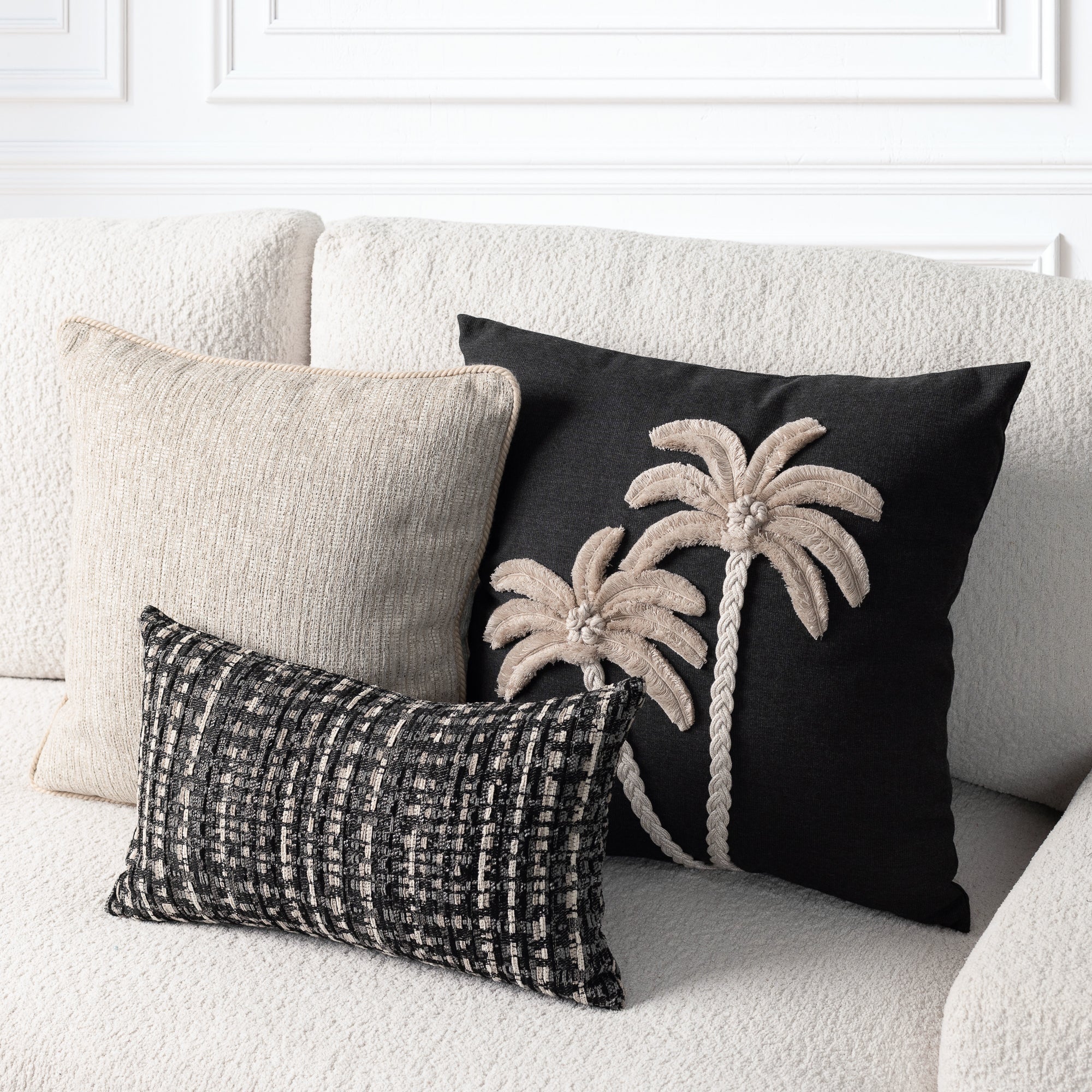 Three decorative throw pillows arranged on a cream textured sofa: a large black pillow with two beige palm tree motifs, a plain beige pillow, and a smaller black-and-beige woven lumbar pillow.